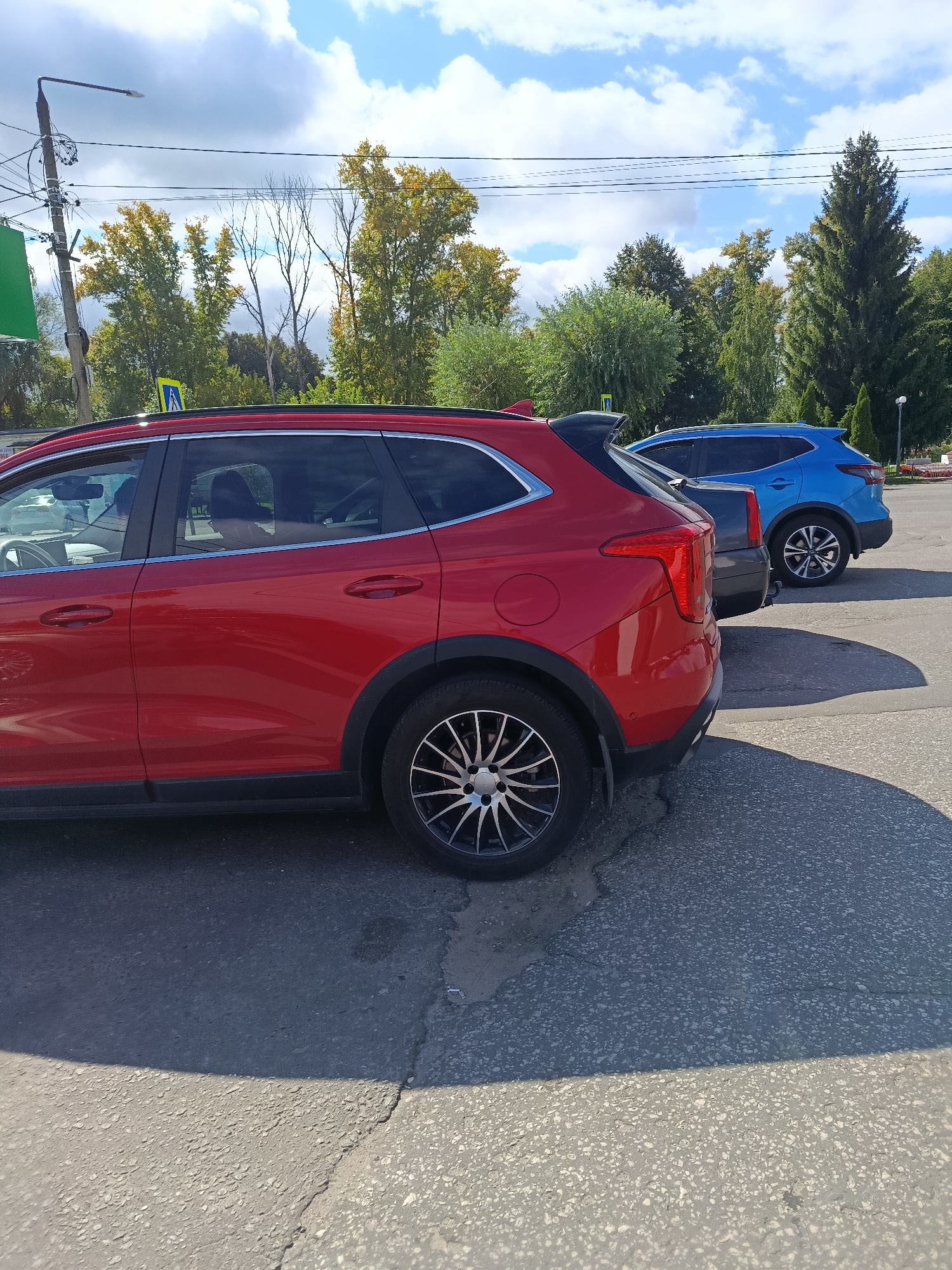 Two cars are parked on an asphalt surface in an outdoor urban environment, possibly a parking lot or street. The foreground car is red, and its right side is prominently visible, showing the rear passenger door, wheel, and part of the front passenger door. The red car has tinted windows and a black spoiler. Behind it, a blue SUV is parked, also seen from its right side. The background features a line of trees with green and yellow foliage, indicating it might be autumn. A utility pole with wires and a street lamp is visible on the left side of the frame. The sky above is blue with scattered white clouds. The scene is illuminated by sunlight, casting shadows on the asphalt. There are no people clearly visible in the image. The location is identified as Verea, Russia.