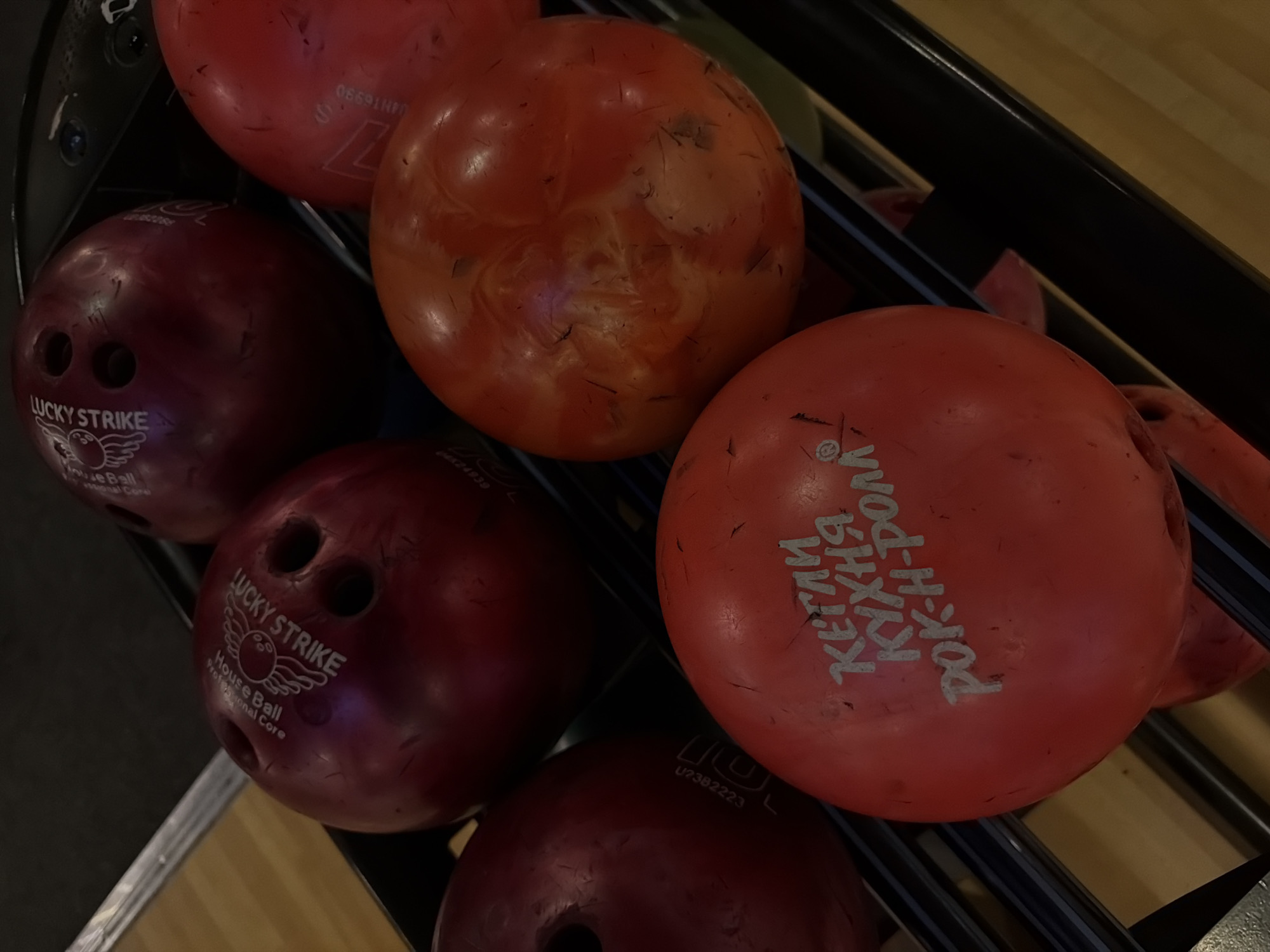 The image shows several bowling balls of varying colors (red, orange, and maroon) stacked in a bowling ball rack. The bowling balls have varying markings and text on them, including "LUCKY STRIKE", "KEГЛИ КУХНЯ РОК-Н-РОЛЛ" (which transliterates to "Kegli, Kitchen Rock 'n' Roll"). Specific serial numbers are visible on some balls. The location Izhevsk, Russia is mentioned in reference to what is said on the ball in Russia.