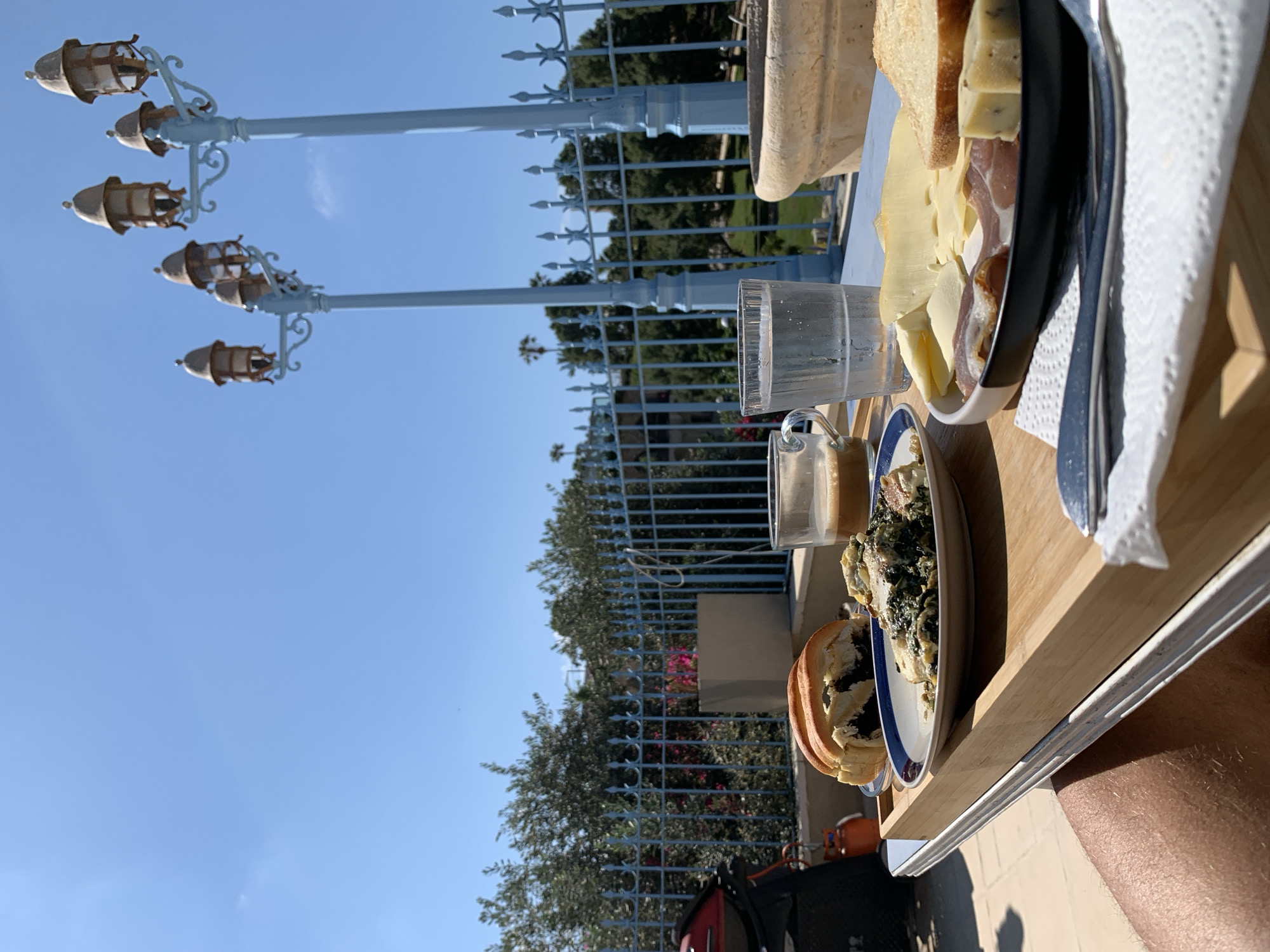 The image shows a meal served outdoors, likely on a balcony or patio, on a wooden tray. There is a plate with cheese and cold cuts, a small glass of a beverage, a plate with some type of stew or vegetable dish and some bread. There is also a knife and paper towels. In the background is a light blue fence and a partially visible light fixture. The clear blue sky suggests a sunny day. The location may be Tel Aviv, Israel, as requested.