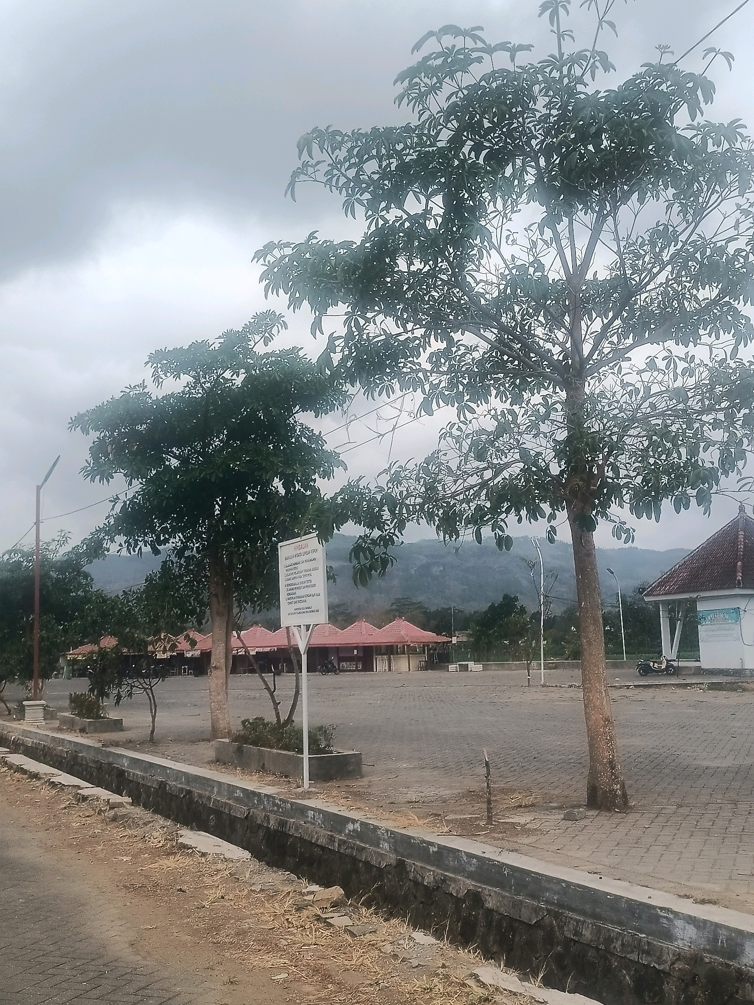 The image depicts an outdoor scene in the city of Gandusari, Indonesia. In the foreground, a concrete drainage ditch runs alongside a paved path. The path is made of interlocking pavers and is bordered by a concrete curb. Beyond the path, a large, open paved area extends, appearing to be a parking lot or public square. Several trees are scattered throughout this area, with one particularly prominent tree in the mid-ground.
In the background, a row of small buildings with red tiled roofs is visible, likely shops or food stalls. Further back, rolling hills covered in vegetation are present under a cloudy sky. On the right side of the image, a white building with a similar red tiled roof stands. A scooter is parked near this building. A white signpost stands in the paved area, displaying text in Indonesian. The overall atmosphere is overcast and appears to be a public area in a tropical or subtropical region. There are no people clearly visible in the image, though the presence of parked vehicles suggests recent activity.