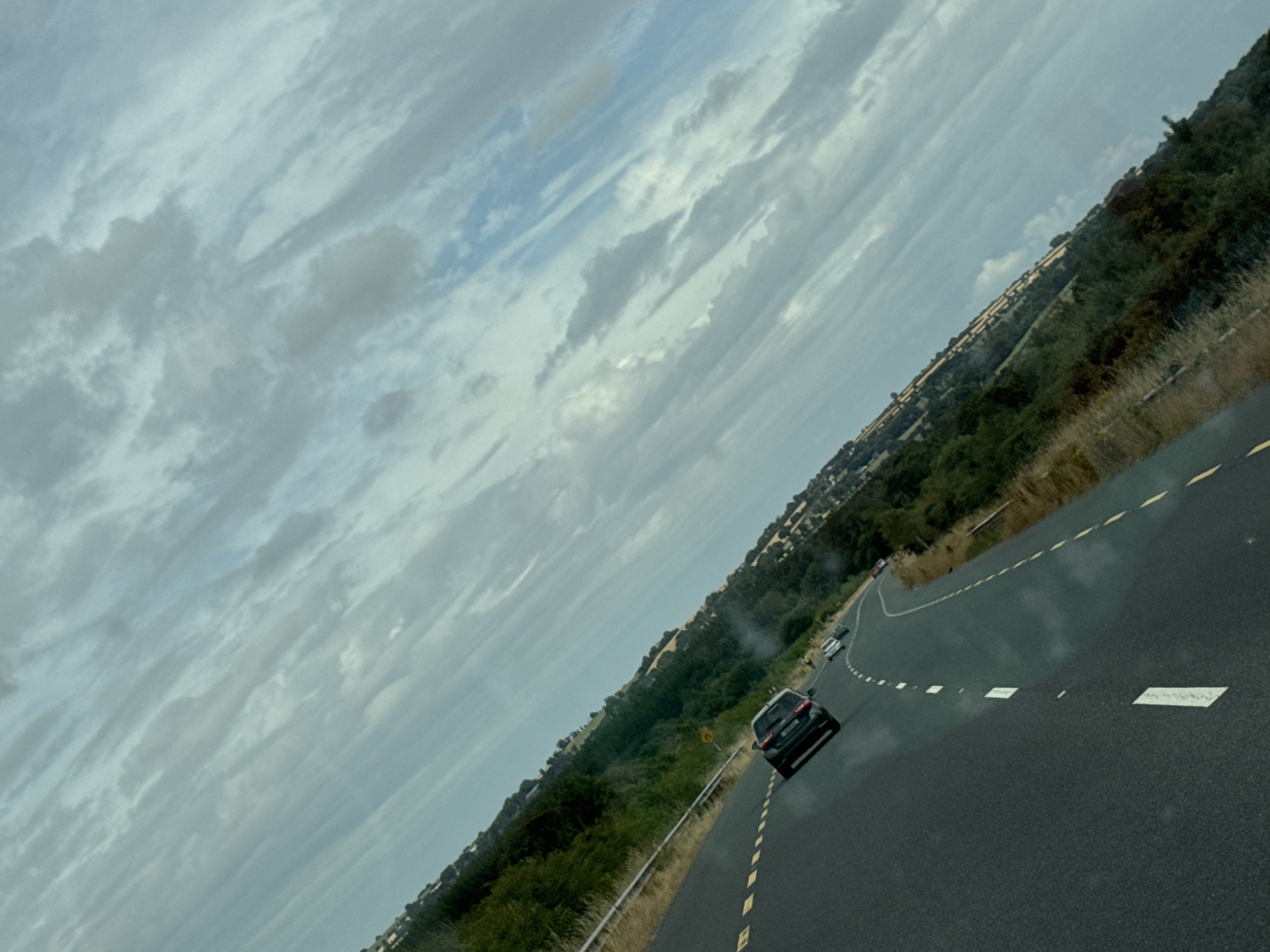 This is a tilted, medium-shot, outdoor, eye-level, horizontal, slightly hazy, natural light, day time photograph taken from a moving vehicle, showing a two-lane road in the countryside. The road is asphalt with a yellow dashed line on the right and a solid yellow line on the left. Several cars are visible on the road ahead. The landscape on both sides of the road consists of rolling hills covered in green grass and scattered trees. The sky is cloudy and appears to be overcast. The image is likely taken in the New Ross Municipal District, Ireland, given the characteristic landscape and vegetation.