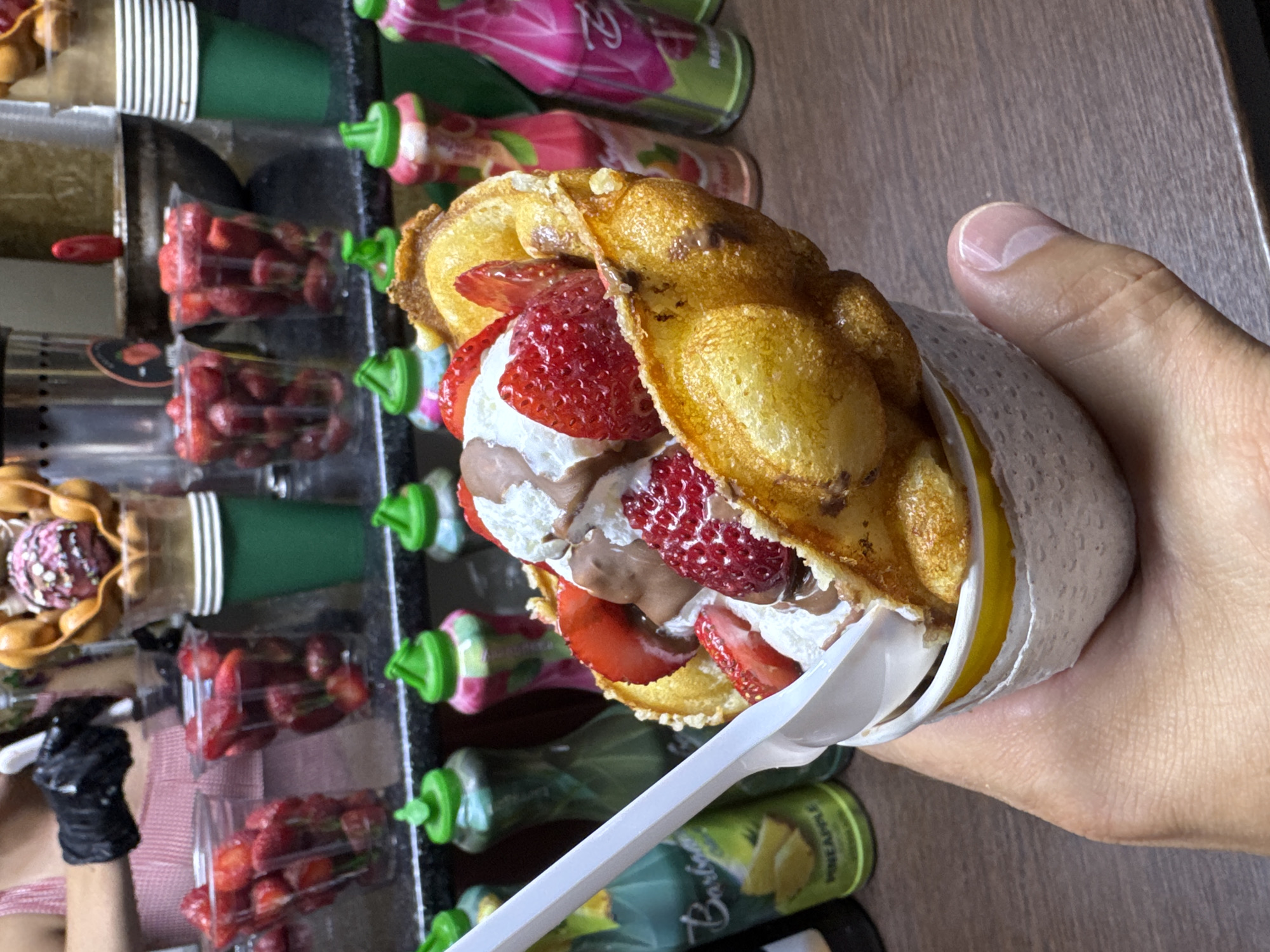 In Zharbulak, Kazakhstan, a person's hand is holding a bubble waffle ice cream cone filled with whipped cream, chocolate sauce, and strawberries. The cone is golden brown and crispy, with a round scoop of ice cream on top. Several bottles of flavored syrups and cups of fresh strawberries are visible in the background, suggesting a dessert stall or shop. A white plastic spoon is inserted into the waffle cone.