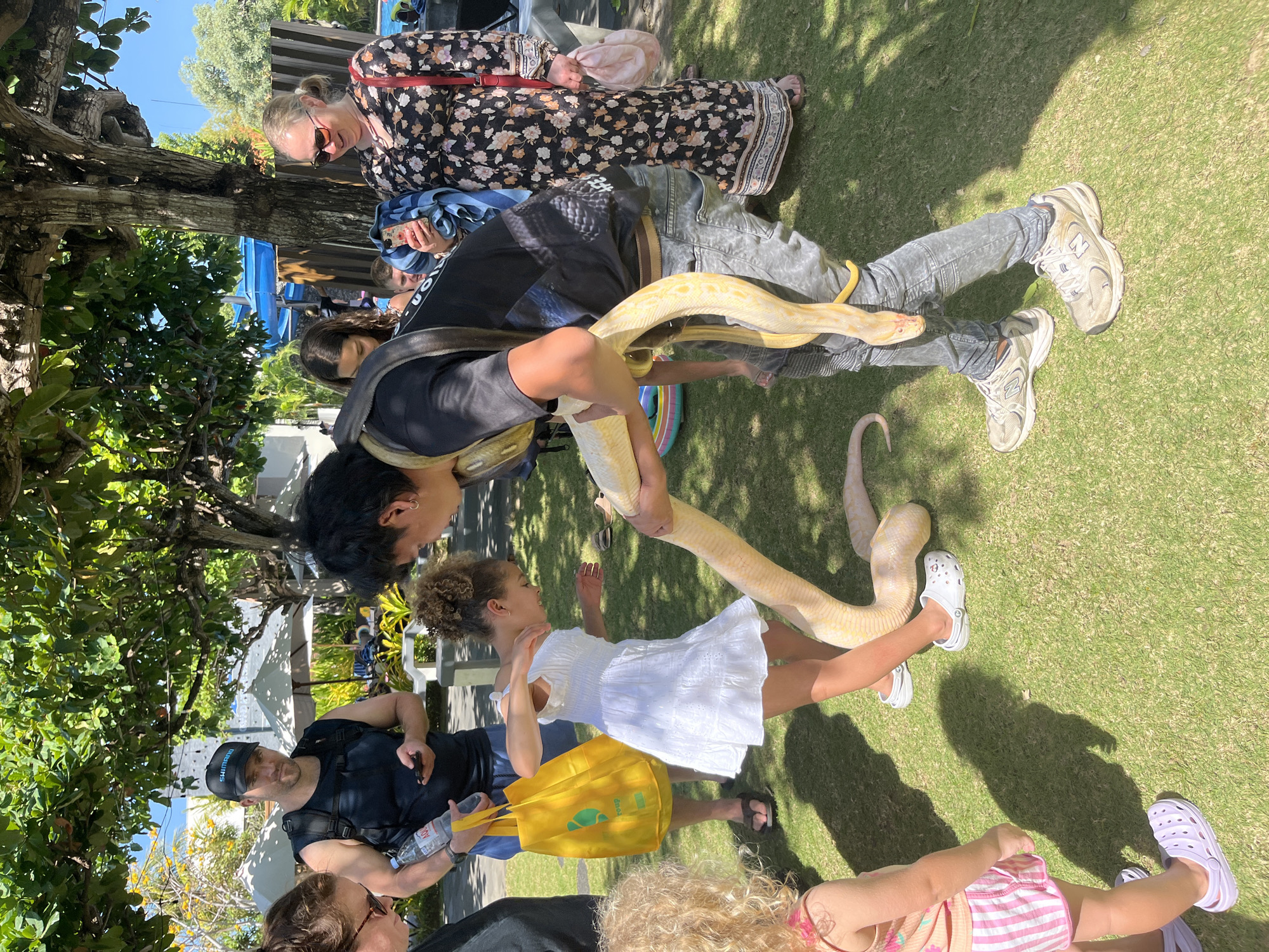 In Kuta Selatan, Indonesia, on a sunny day, a group of people gather on a grassy lawn under the shade of trees. A young girl in a white dress is interacting with a large, pale yellow python being held by a young man. The man, wearing a black t-shirt, grey jeans, and sneakers, has the snake draped around his neck and is holding it with both hands, bringing it closer to the girl. The girl, with curly hair, appears engaged and curious as she reaches out towards the snake. To the left, a toddler in a striped outfit and purple crocs stands with her back to the camera, looking towards the snake. A man wearing a black tank top and a baseball cap stands behind the girl, holding a yellow bag and a bottle of water, while looking towards the interaction. Another woman with blonde hair and sunglasses is also present, looking on. In the background, umbrellas, a blue water slide, and buildings suggest a resort or recreational area. The overall scene captures a moment of interaction with wildlife in an outdoor setting.