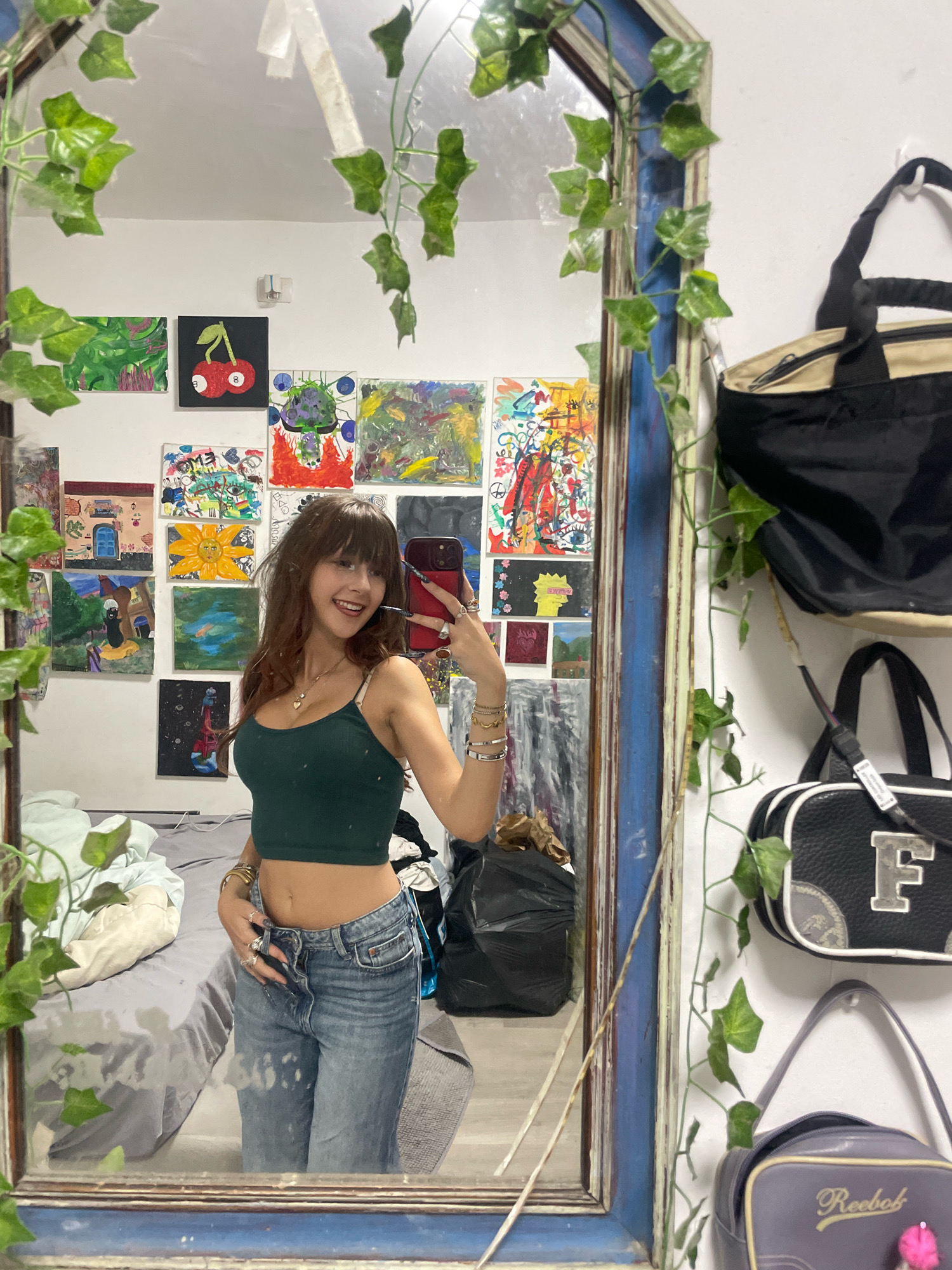 A young woman with brown hair and bangs, wearing a green crop top and blue jeans, poses for a mirror selfie in a room. She has on multiple rings and bracelets and is holding a red smartphone. The room has white walls covered with various paintings and artificial ivy vines draped around a blue-trimmed mirror. A bed with gray bedding is visible behind her in the reflection, along with several bags hanging on the wall. The scene appears to be indoors, possibly in a bedroom or studio. The location of Raanana, Israel is not discernible from the image.