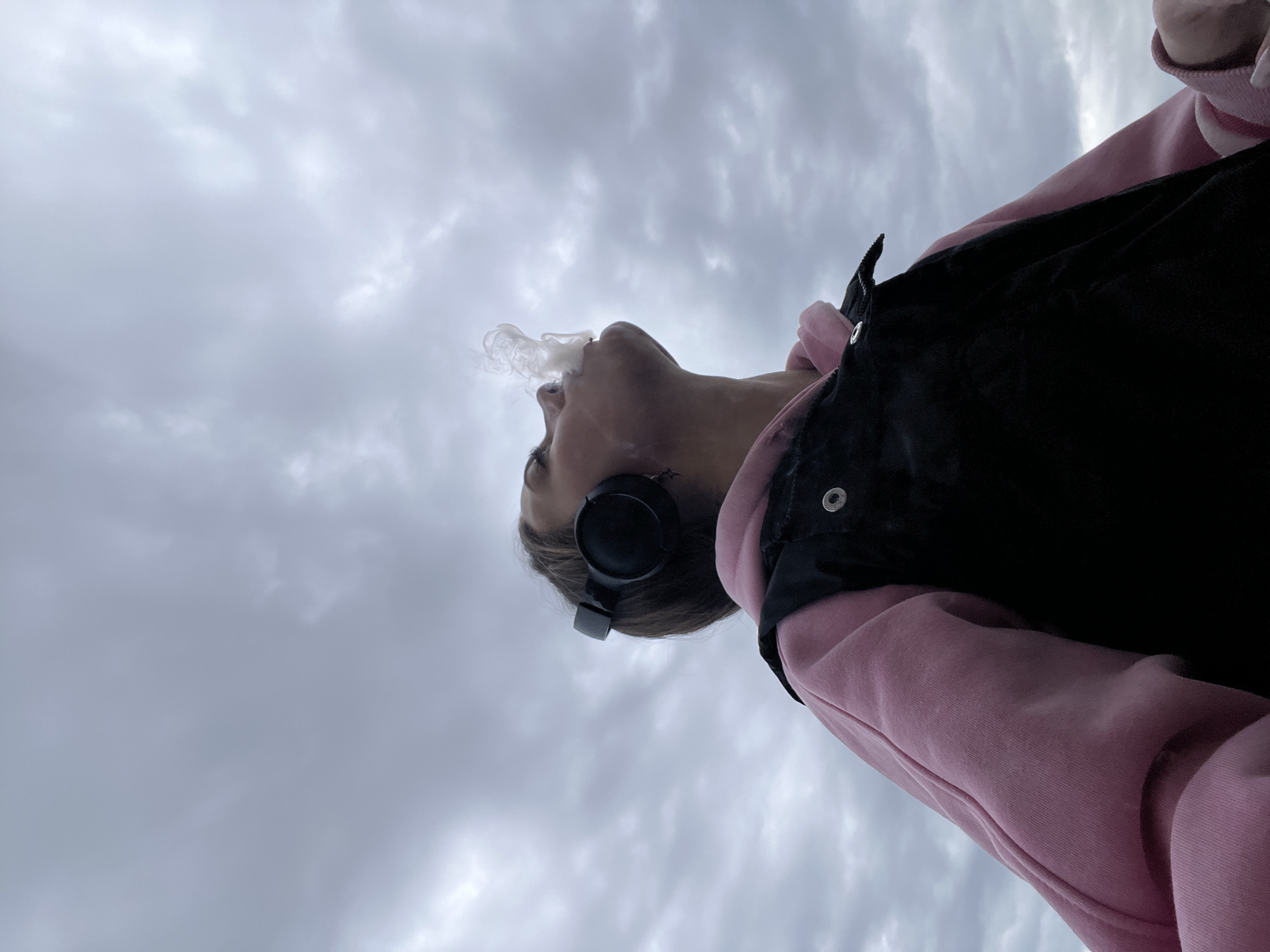 A low-angle, upward-facing shot reveals a person with their head tilted back, exhaling vapor into the sky. The person is wearing headphones around their neck and a black jacket with a pink hoodie underneath. The sky is overcast with grey clouds. The precise location in the city of Фоки, Russia is indeterminable.