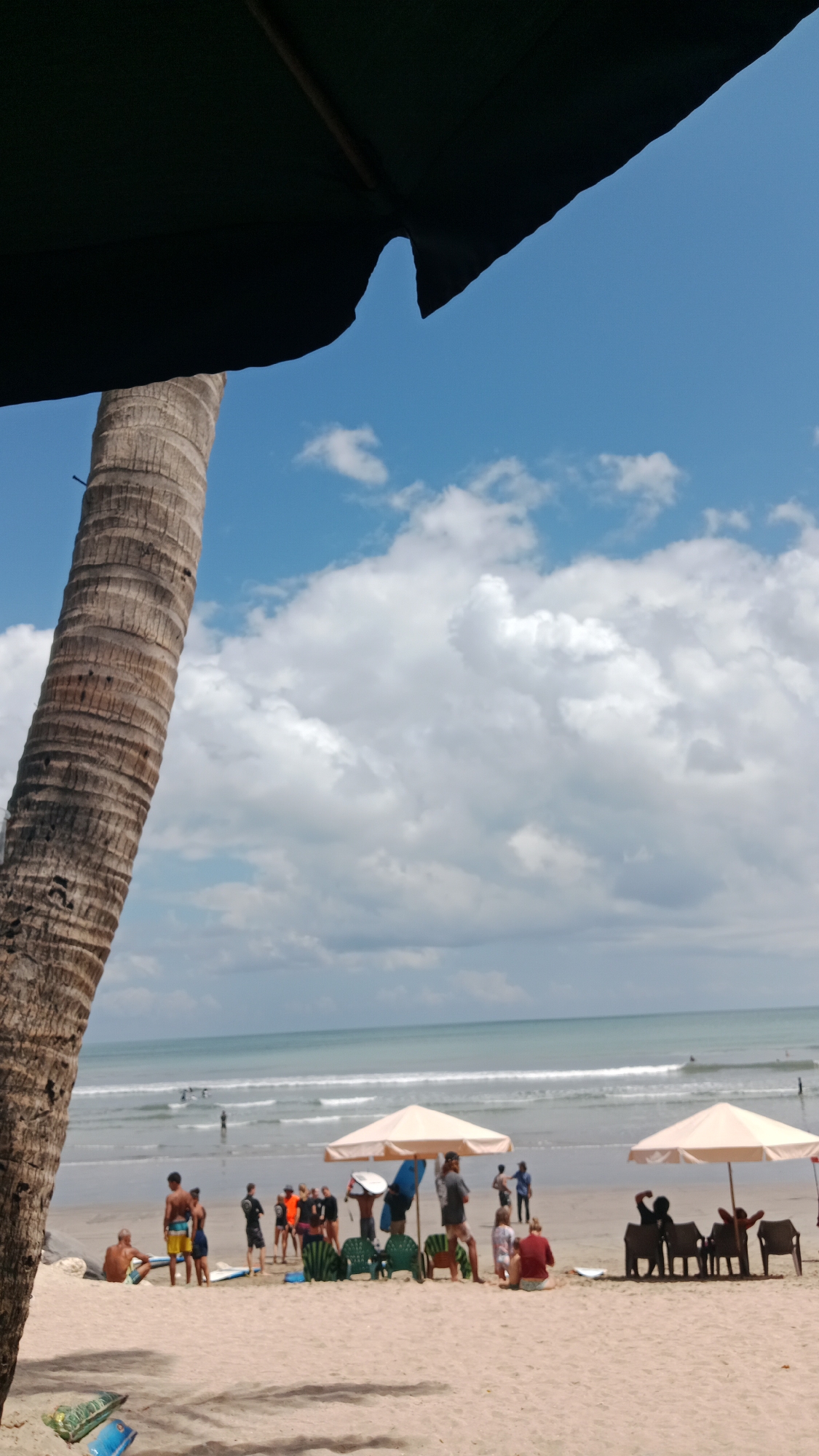 In Kuta, Indonesia, a beach scene unfolds with a towering palm tree trunk in the foreground, partially obscuring a bright blue sky dotted with fluffy white clouds. The ocean stretches out to the horizon, with gentle waves breaking on the shore. A sandy beach is populated with numerous individuals. Some are gathered near beach umbrellas and chairs, while others are seen walking, standing, or sitting on the sand. Several surfboards are visible, indicating recreational activities. In the background, more people are in the water, some appearing to be surfing. The overall atmosphere suggests a warm, sunny day at a popular tourist destination.
