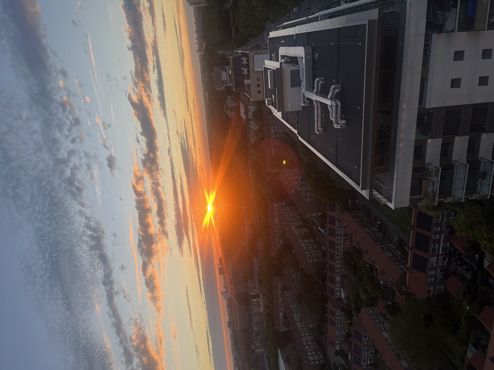 The image captures a vibrant sunset over a cityscape, likely in an urban setting in Unknown, Unknown. The sun, a brilliant orange disc, is positioned low in the sky, casting long rays that create a starburst effect. Wispy, elongated clouds are illuminated with shades of orange, pink, and yellow, contrasting with the pale blue of the upper sky. Below the sky, a dense collection of buildings, characterized by red-tiled roofs, stretches across the mid-ground. These residential structures are interspersed with trees, creating a textured urban landscape. In the foreground, a modern building with a flat, dark roof dominates the right side of the frame. This building features prominent external ductwork and a white facade with multiple windows and balconies. The overall impression is an aerial view of a city during the golden hour, with a blend of residential and commercial architecture under a dramatic sky.