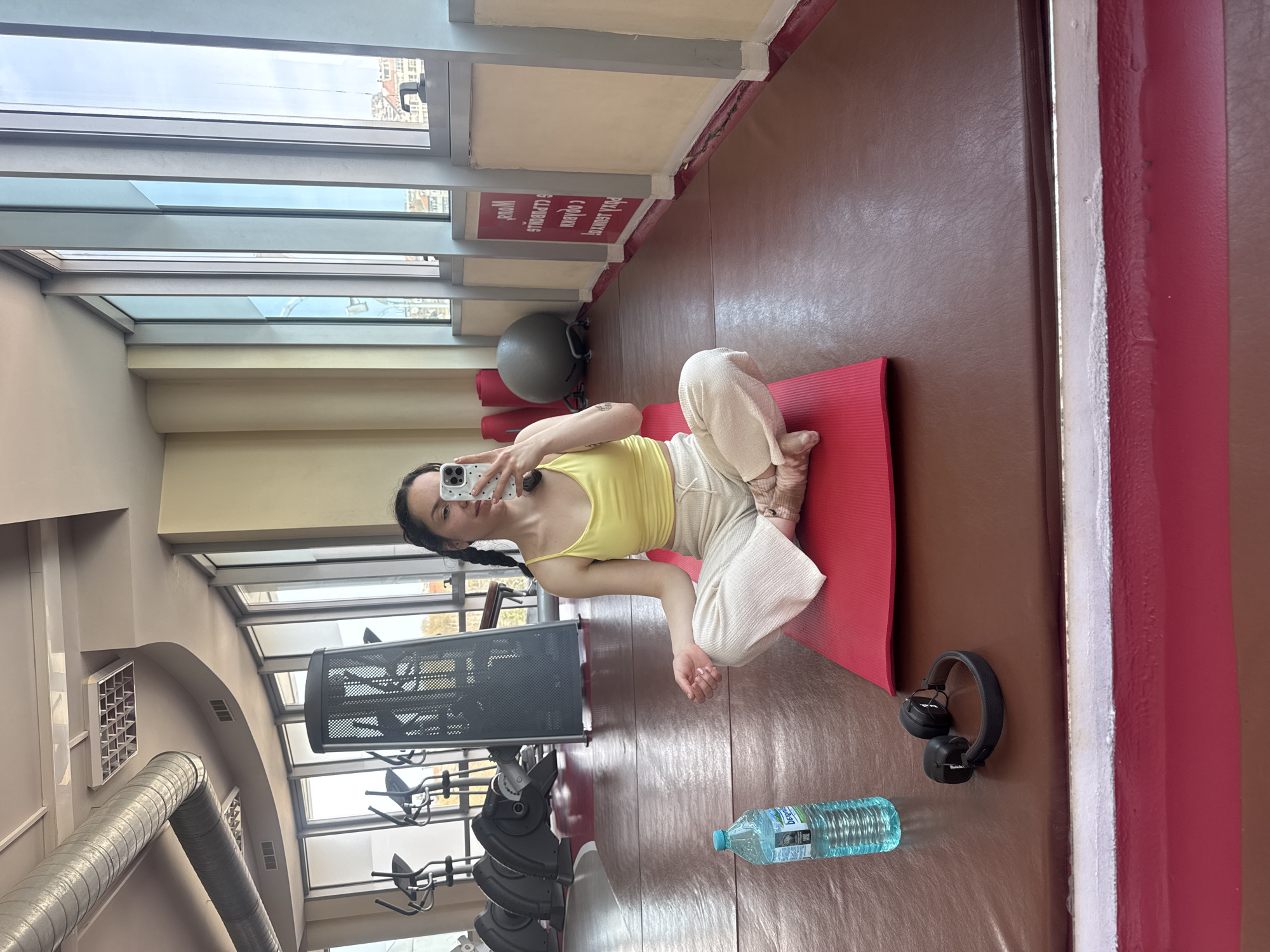 A young woman is sitting cross-legged on a red yoga mat in what appears to be a gym or fitness studio. She is holding a cell phone up to take a picture of herself. She has dark hair in braids and is wearing a yellow tank top and light-colored pants. A water bottle and black headphones are on the floor next to the mat. Fitness equipment, including elliptical machines and a weight rack, are visible in the background. Tall windows stretch across the left side of the room, offering a glimpse of buildings outside. Based on available visual information and given the context of "Burgas, Bulgaria," it is not possible to confirm the exact location.