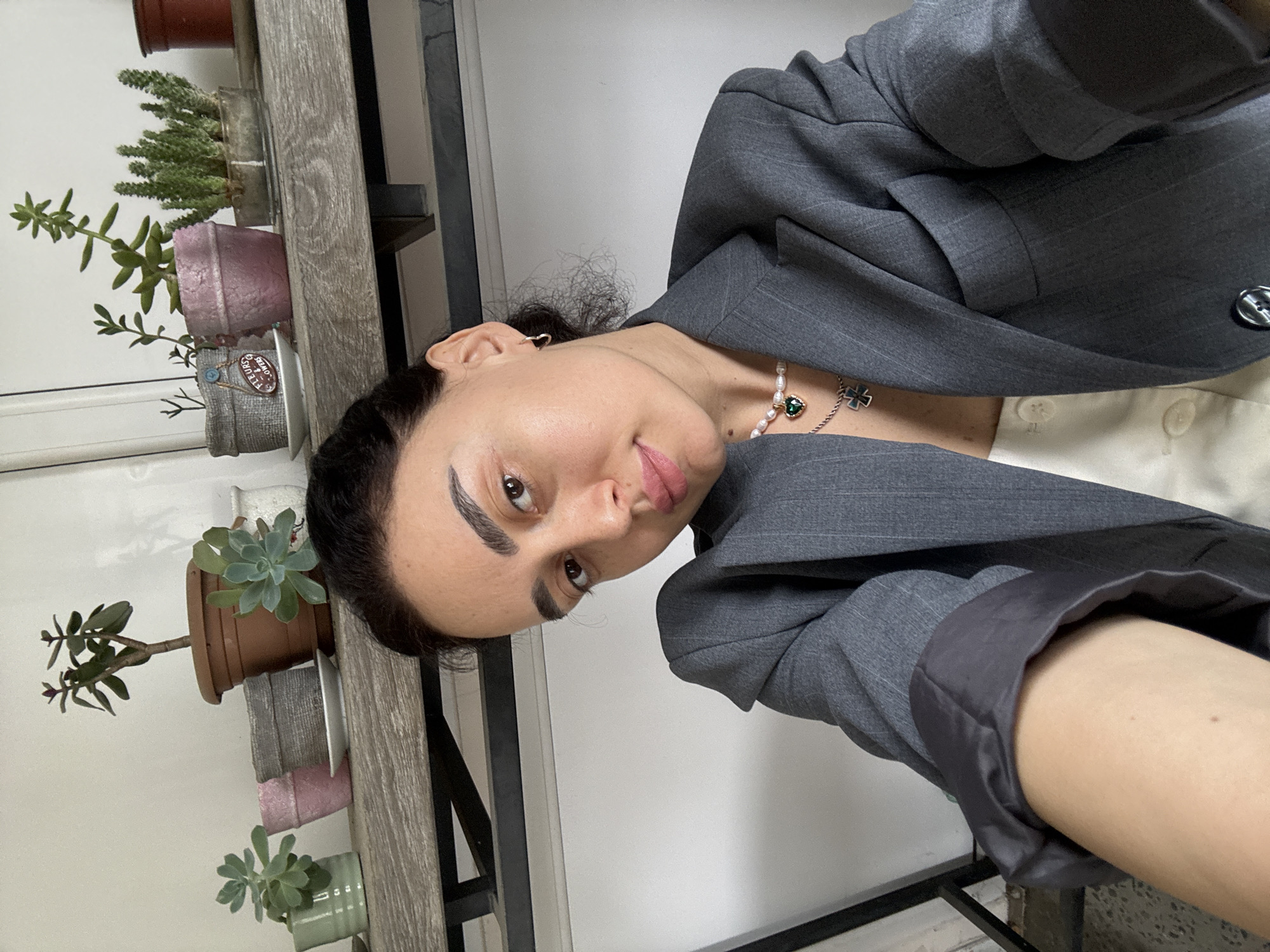 A woman poses for a selfie indoors. She is wearing a grey blazer over a white button-down shirt and a necklace. Behind her is a wooden shelf with various potted succulents. The location cannot be determined to be Tbilisi, Georgia from the visual information provided.