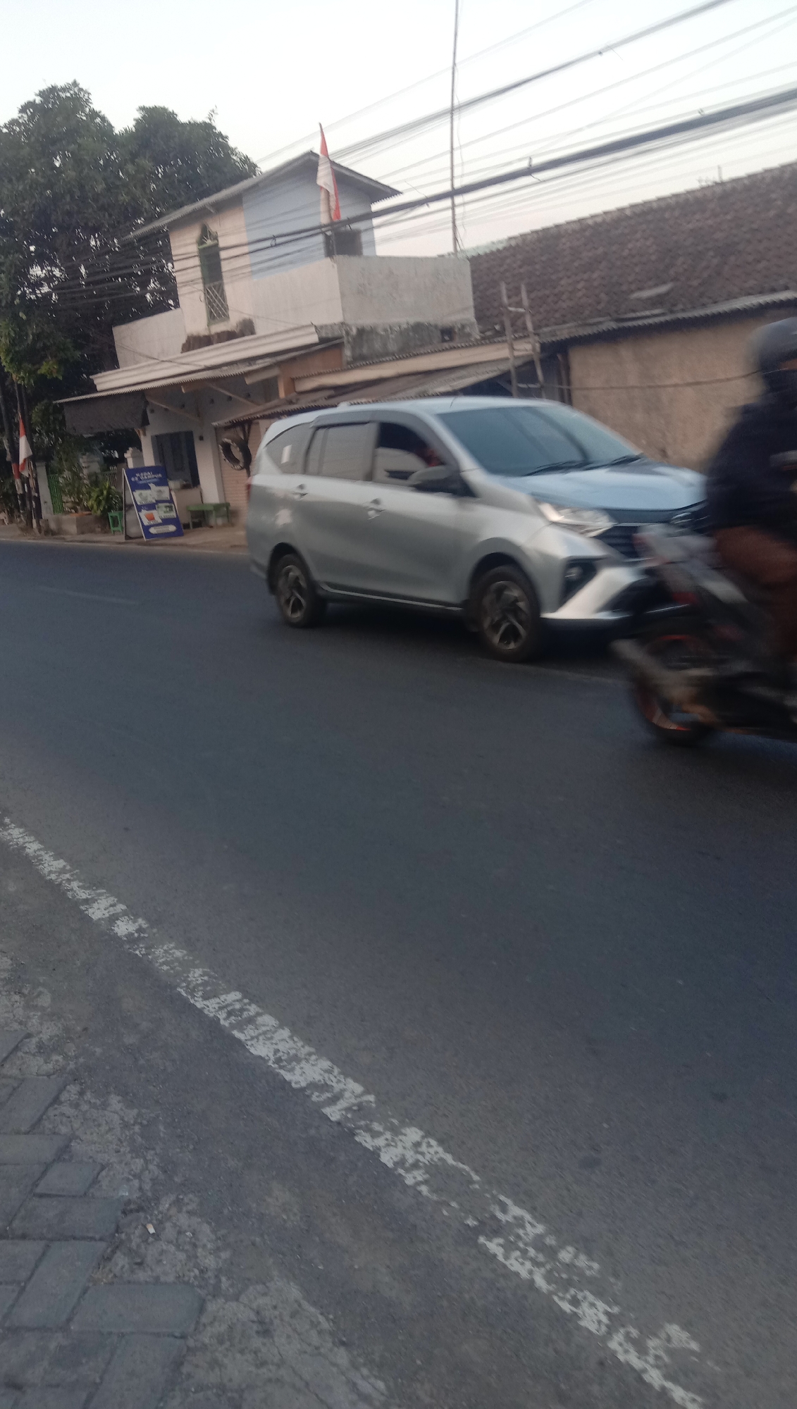 The image depicts a street scene in Karanganyar, Indonesia, with a silver MPV (multi-purpose vehicle) driving on the right side of the road, facing left. A motorcycle is passing the MPV on its right. The road is paved with asphalt and has a white dashed line on the left side. The foreground shows a section of paved sidewalk or walkway with rectangular stones. In the background, there are buildings, including a two-story structure with a white and light blue facade and an Indonesian flag on a pole. Trees with green foliage are visible to the left of the building. Power lines are strung across the sky. The scene appears to be illuminated by natural daylight, possibly during late afternoon or early morning given the soft lighting.