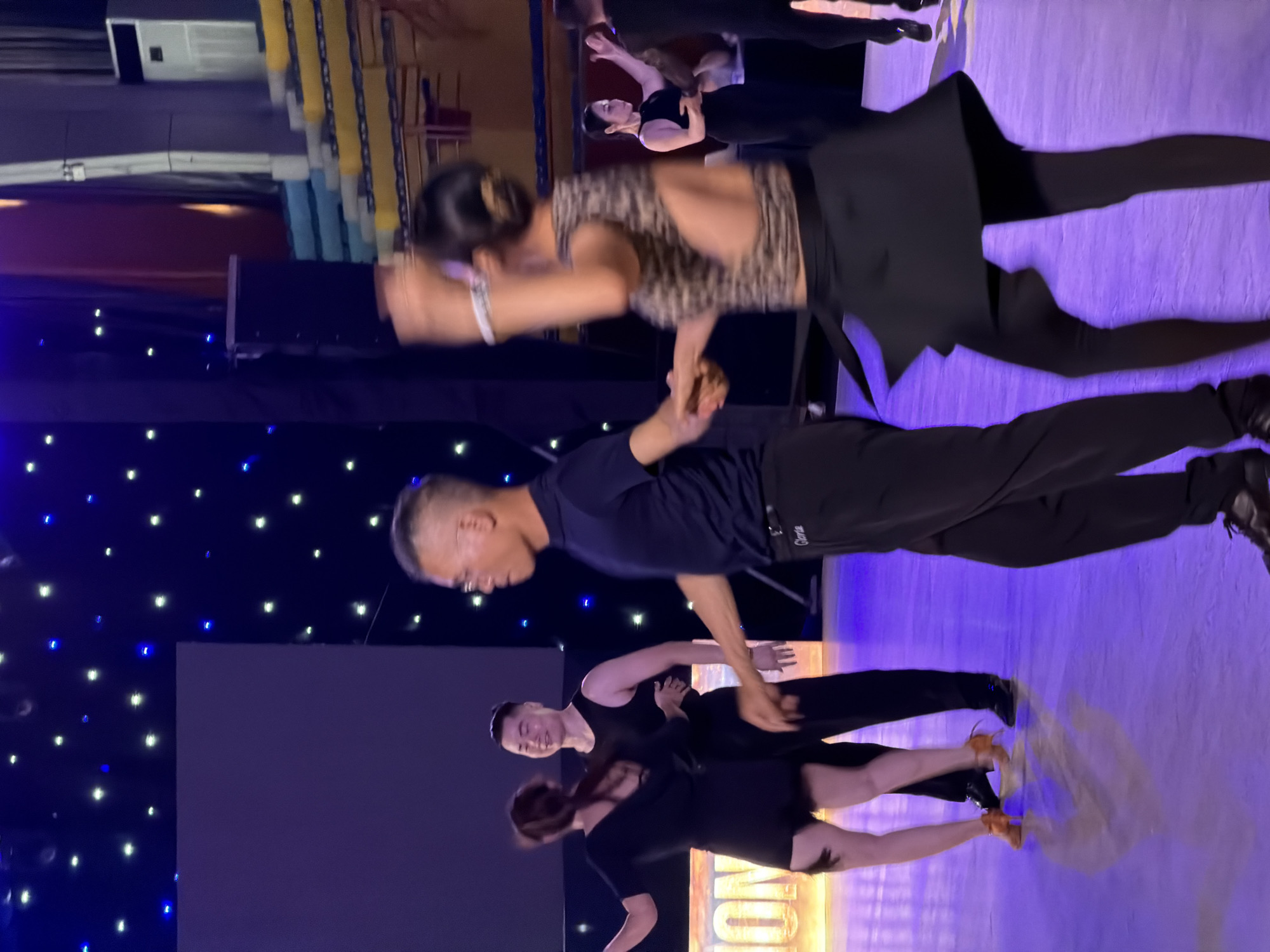 The image depicts a ballroom dance competition or performance taking place indoors. The scene is illuminated by stage lighting, with a dark blue backdrop adorned with numerous small, white and blue lights, resembling stars. In the foreground, several couples are engaged in dancing. One couple in the center is dressed in black attire, with the male dancer wearing a sleeveless shirt and the female dancer in a short black dress with fringe detailing on the hem. Another couple is visible to the right, with the female dancer wearing a leopard-print top and a black skirt, and the male dancer in black attire. A third couple is to the left, also in black. The floor is a reflective purple surface, suggesting it is a dance floor. The overall atmosphere is one of performance and movement. While the specific location within Hanoi, Vietnam, cannot be determined from the image alone, the presence of dancers and stage setup indicates a venue dedicated to artistic performances or competitions, such as a cultural center or a performance hall.