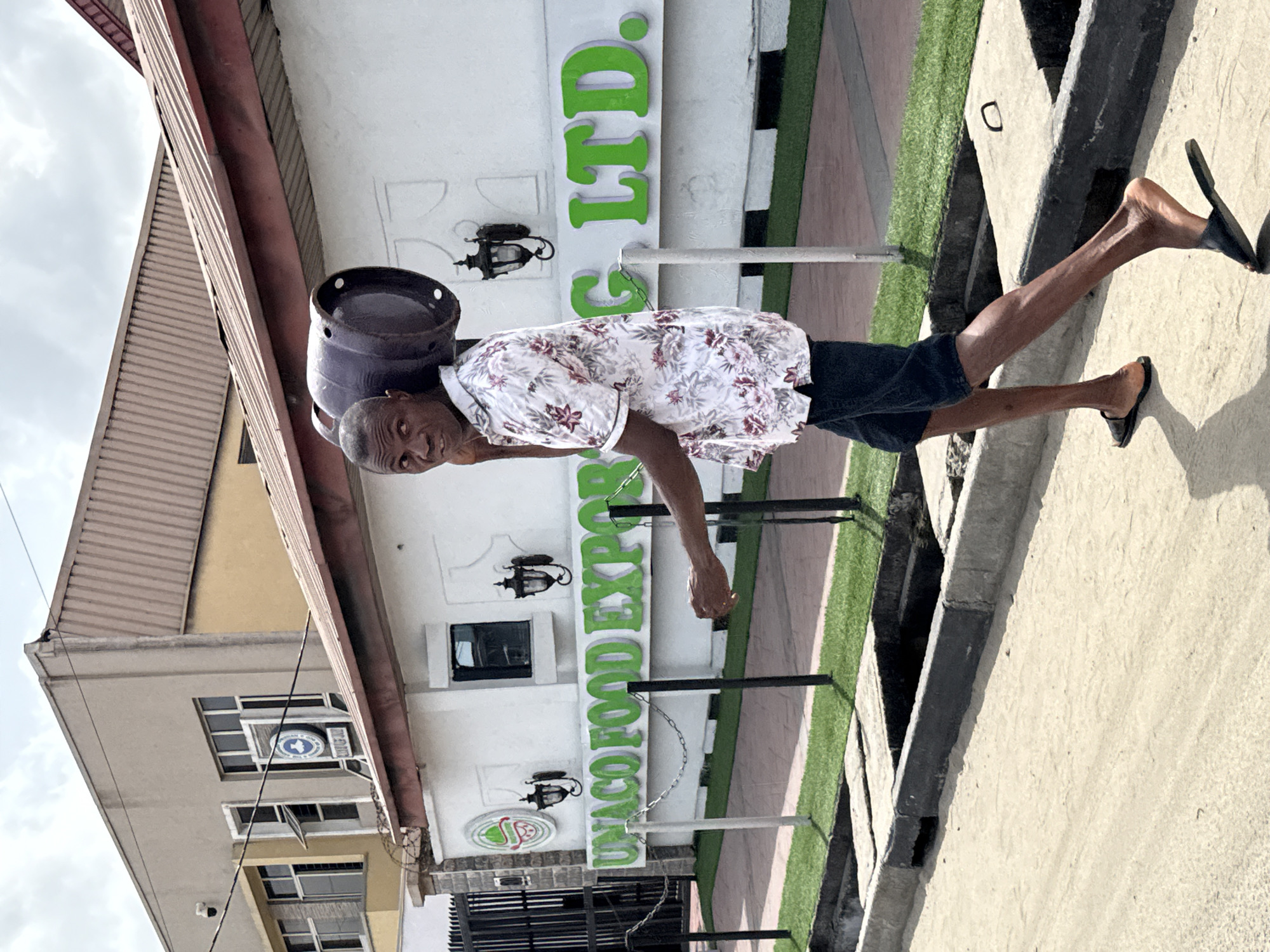 The image, taken in an urban or commercial setting, features a man carrying a dark-colored gas cylinder balanced on his head as he walks. He is wearing a short-sleeved, collared shirt with a white background and a floral pattern in red/maroon and grey, paired with dark shorts and dark flip-flops. The man has short, dark hair and dark skin, and he looks towards the viewer with a neutral expression.
Behind the man, a light-colored building with a brown roof is visible. Large green letters on its facade spell "G LTD." at the top, and below that, "UNACO FOOD EXPO". The building also features decorative black sconce lights. To the left, another building with multiple windows displays a circular blue sign that appears to be a bank logo with an arrow and the words "BANK OF..." partially visible.
The ground is a mix of concrete pavement and a strip of artificial green grass bordering the main building. A concrete curb or drain structure runs parallel to the walking path on the right side of the image. The sky is partly cloudy, suggesting it is daytime. The scene reflects a common method of transportation in many regions, including Ado, Nigeria, where the image was captured.