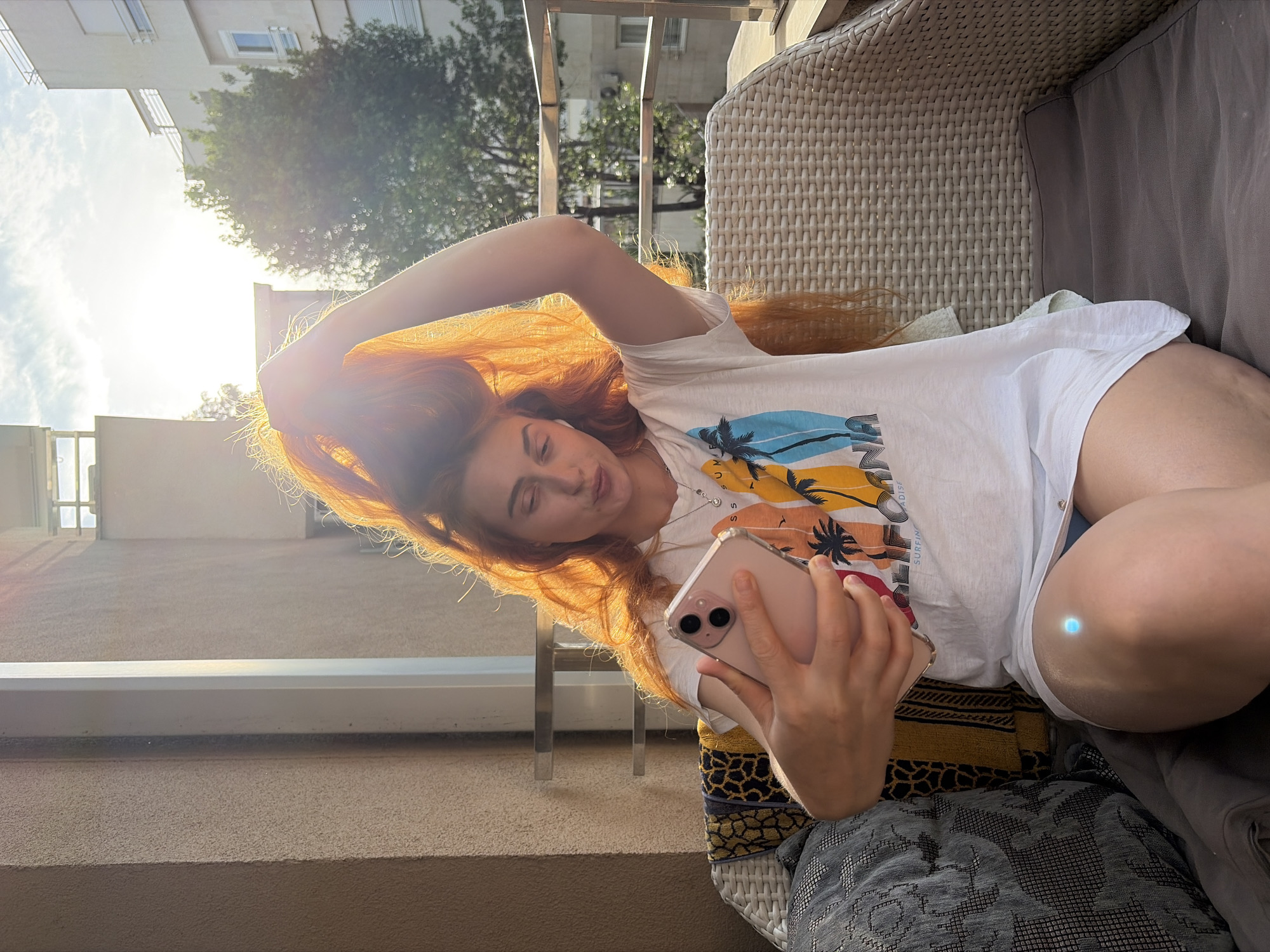 A young, fair-skinned woman with long, auburn-red hair is sitting and taking a selfie on a balcony in what appears to be Budva, Montenegro. The woman is angled to the side, wearing a white t-shirt with a colorful graphic print, and is holding a pink iPhone. Her left arm is raised and her hand is holding her hair. She is seated in a beige woven chair with gray cushions and a patterned decorative pillow. The balcony has a light-colored stone floor and a metal railing. The background shows buildings, trees, and the sunny sky typical of the Budva region.