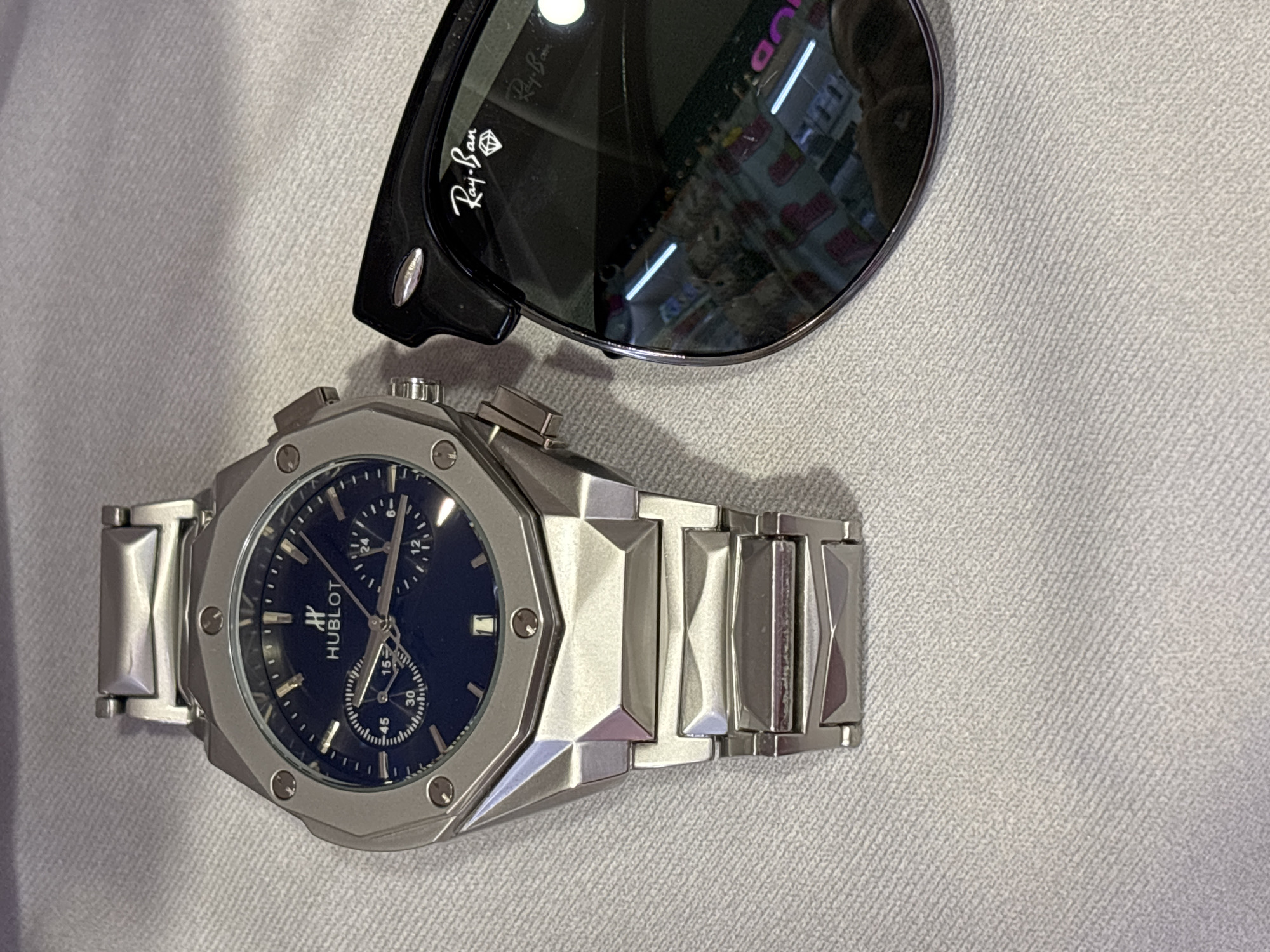 The image depicts a silver Hublot watch with a navy blue face resting next to a pair of black Ray-Ban sunglasses on a light gray textured surface. The watch features a hexagonal bezel with visible screws and a metal link bracelet. The sunglasses are partially visible, with the Ray-Ban logo displayed on the lens. The reflection in the sunglasses suggests the interior of a store, potentially in Saint Petersburg, Russia, although the exact location cannot be definitively determined.