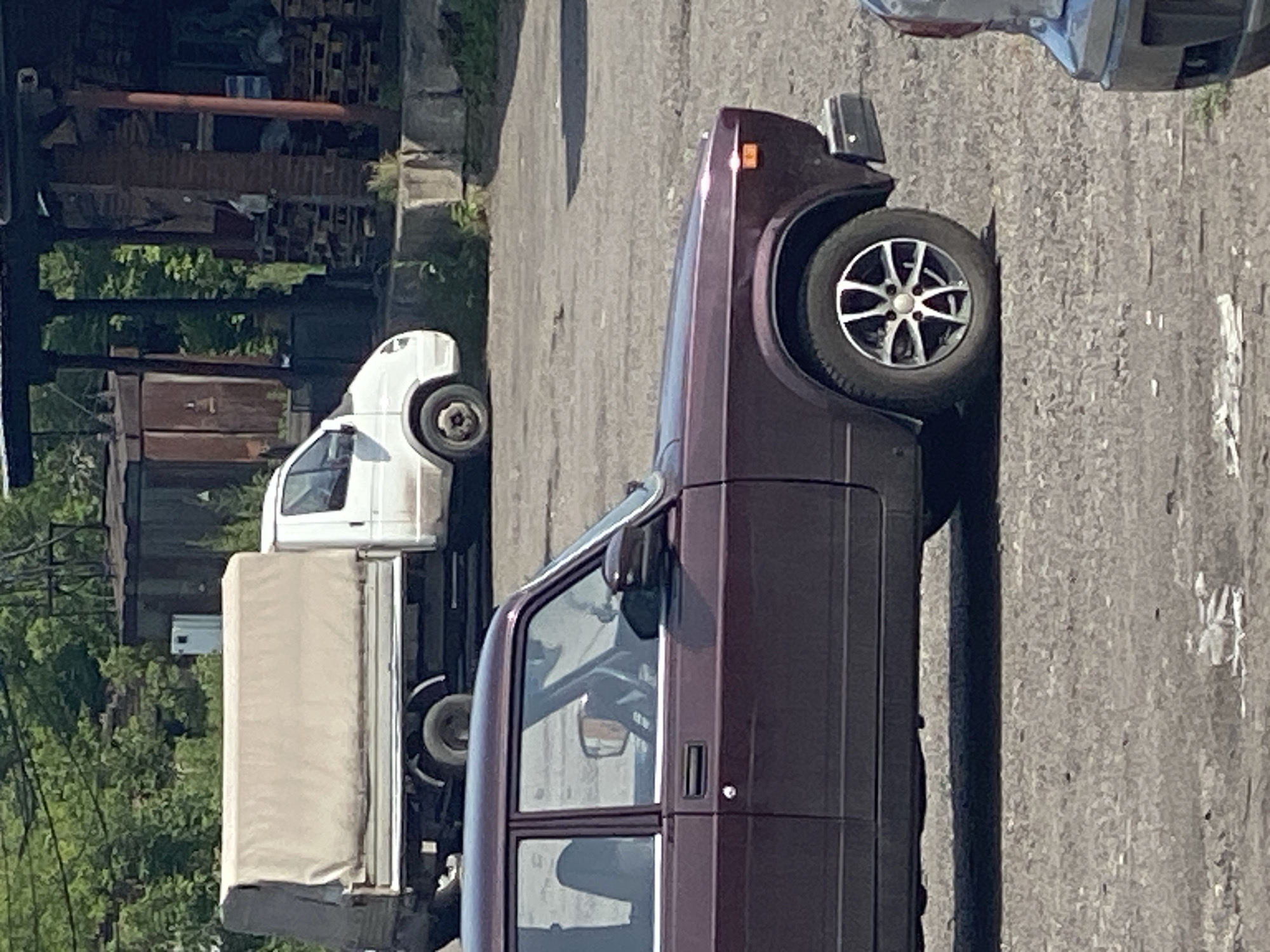 A mid-shot, eye-level, slightly tilted outdoor photograph depicts a scene likely in Novokuznetsk, Russia. In the foreground, a dark red sedan is parked on a gravel lot, its right side visible. The car's wheel, with a silver multi-spoke rim, is clearly visible. In the background, a white truck with a beige canvas cover over its bed is also parked. Behind the truck, a weathered building or structure with wooden elements and stacked pallets is partially visible. The environment appears industrial or utilitarian, with trees in the distance. No people are present in the frame.
