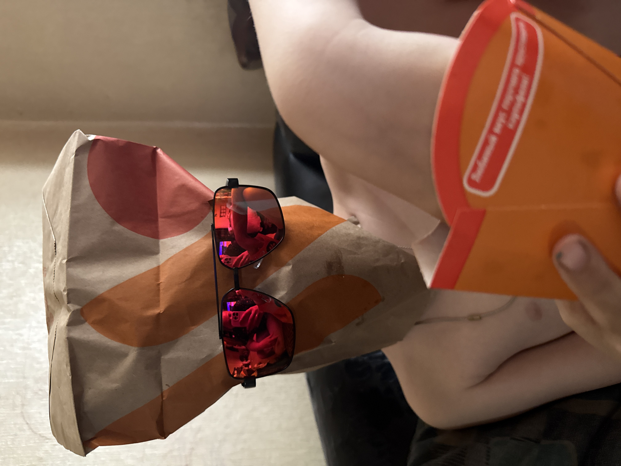 An indoor, close-up shot captures a person with light skin, partially visible from the chest up, holding a fast-food container. The individual's right arm and shoulder are visible, along with a thin chain necklace around their neck. The person is dressed in dark fabric, possibly camouflage pants, visible in the lower portion of the image.
In the foreground, a brown paper bag, likely from McDonald's given the visible orange and red semi-circle logo, holds red mirrored sunglasses. The sunglasses reflect distorted images of an interior space, including bright light sources and vague shapes. Adjacent to the paper bag, the person holds an orange cardboard container, consistent with a McDonald's fries package.
Visible text on the orange container reads "Большой вкусный перекус!" (Bolshoy vkusnyy perekus!), which translates to "Big tasty snack!" or "Big delicious snack!", indicating Russian language branding. The activity appears to be the consumption of fast food. The setting is an unidentifiable indoor room with a light-colored background. The image is taken in Dmitrov, Russia.
