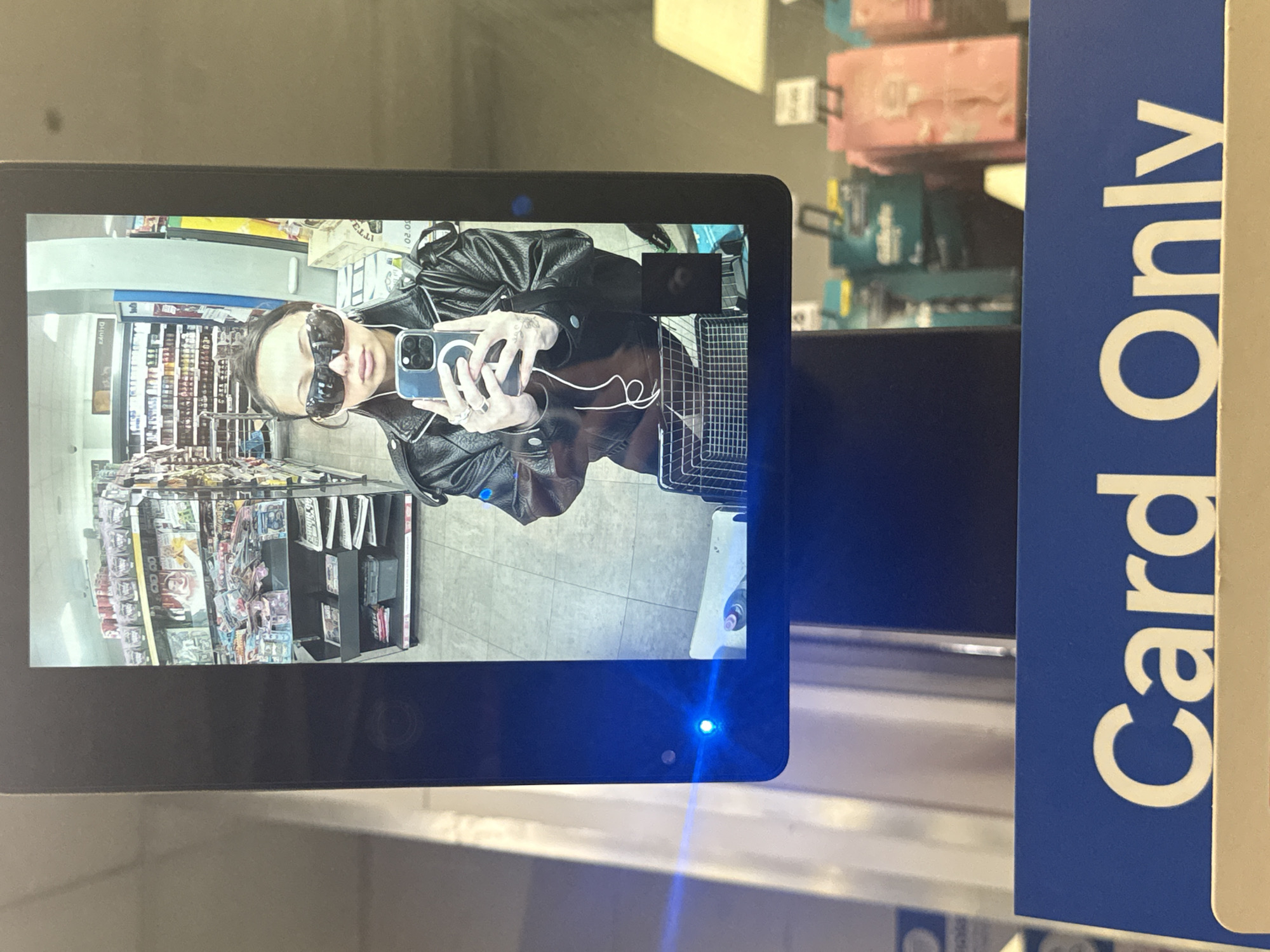 This image captures a person taking a selfie reflection in a vertical digital screen within a retail environment. The image itself is rotated 90 degrees clockwise, but when mentally re-oriented, the screen and its contents are upright.
The subject, appearing to be a woman, is reflected on the screen from approximately the chest up. She wears dark, oversized sunglasses, large hoop earrings, and a black leather jacket. Several rings adorn her fingers as she holds an iPhone, identifiable by its multiple camera lenses, pointed towards the screen to capture her reflection.
The background, visible in the screen's reflection, shows an aisle of a store with various products displayed on shelves, including packaged items and magazines. The floor appears to be tiled. A small blue indicator light is visible on the bottom right corner of the screen, indicating it is powered on and likely active.
To the right of the screen, a prominent blue sign with white text clearly reads "Card Only," suggesting the location is a self-checkout or payment station in a store. The setting is an indoor commercial space, likely a supermarket or convenience store. The lighting is artificial and bright. The subject's expression is obscured by the sunglasses.