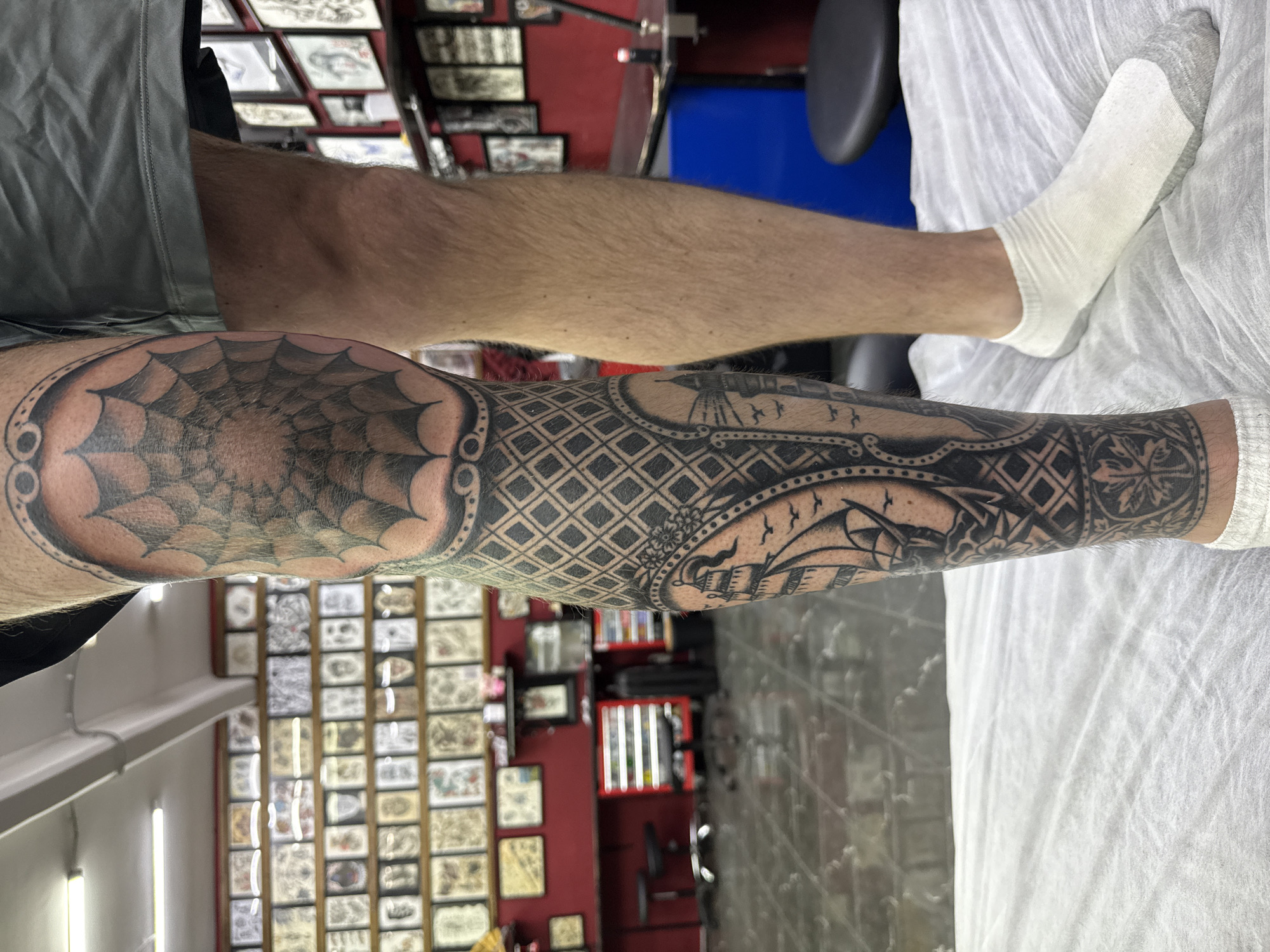 A person with a full leg tattoo is seated. The tattoo features a spiderweb on the knee, geometric patterns mid-leg, and a sailboat design towards the ankle. The person is wearing white socks and grey shorts. The scene appears to be inside a tattoo studio, with framed tattoo designs on the wall. Tattoo equipment is visible in the background, and a blue chair is also present. The location is not identifiable as Moscow, Russia from the image.