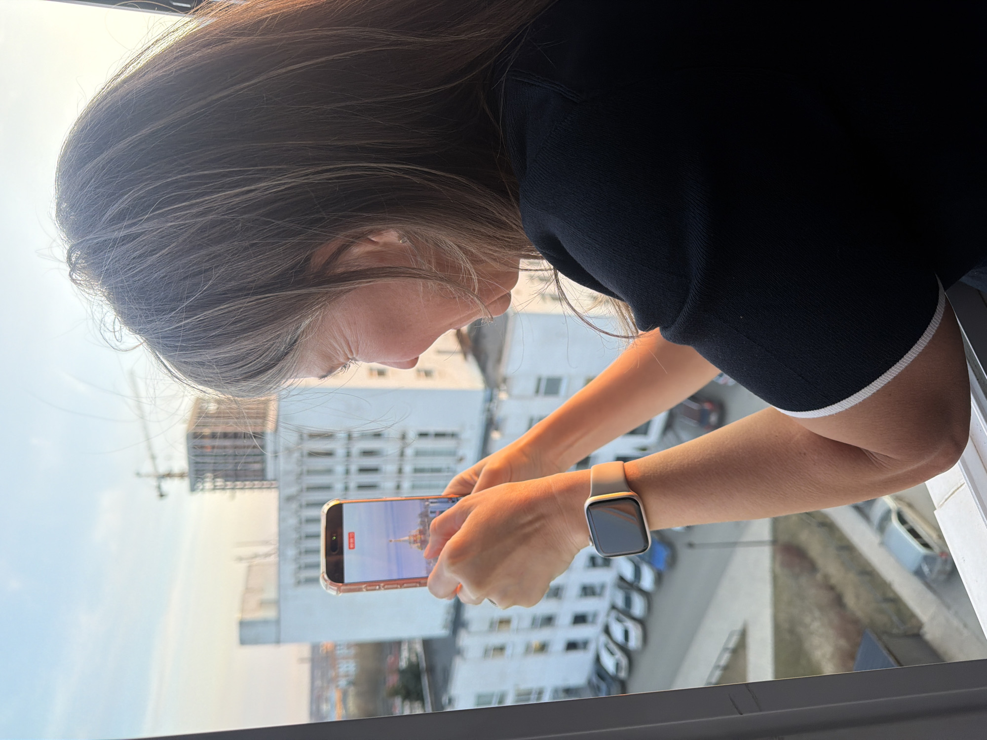 A woman is taking a video with her phone from a high vantage point, likely a balcony or window. The phone screen shows a building with a golden dome, which could be the Alexander Nevsky Cathedral in Izhevsk, Russia. She is wearing a black shirt with white trim on the sleeves and an Apple Watch on her left wrist. In the background, there are other buildings, a construction crane, parked cars, and a clear sky.