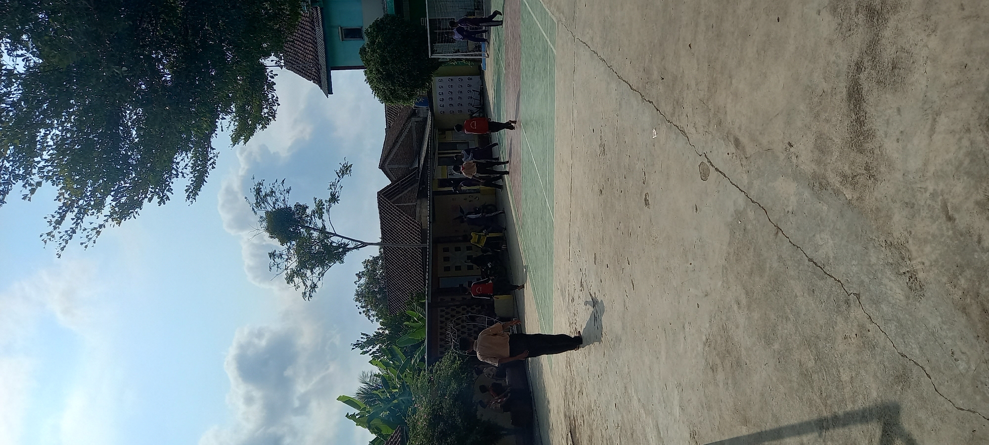 A bright, partially cloudy sky looms over a schoolyard in Cintaratu, Indonesia. In the foreground, a cracked concrete courtyard is partially visible, with a shadow cast from an unseen object on the left. In the mid-ground, a group of children in uniforms are scattered across a multi-colored court, which appears to be marked for badminton or volleyball. Some children are walking, while others are standing or appear to be playing. Behind the court, a low-slung building with a tiled roof is visible, with trees and foliage surrounding it. Further back, a taller, light blue building with a darker roof stands out against the sky. The scene depicts a typical day at a school in a tropical climate, with children engaged in outdoor activities.