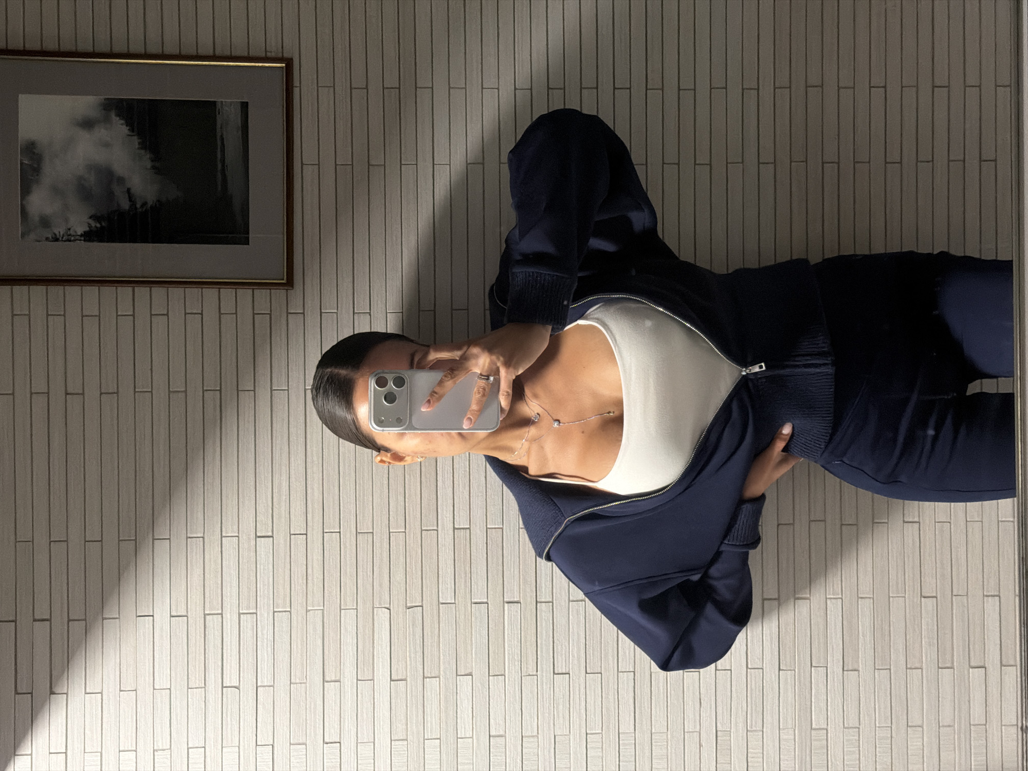A person is taking a mirror selfie. They are wearing a white top and a dark blue zip-up jacket. A framed picture hangs on the wall behind them. The location is a room with off-white, rectangular tiled walls. There is no indication that the location is Krasnoyarsk, Russia.
