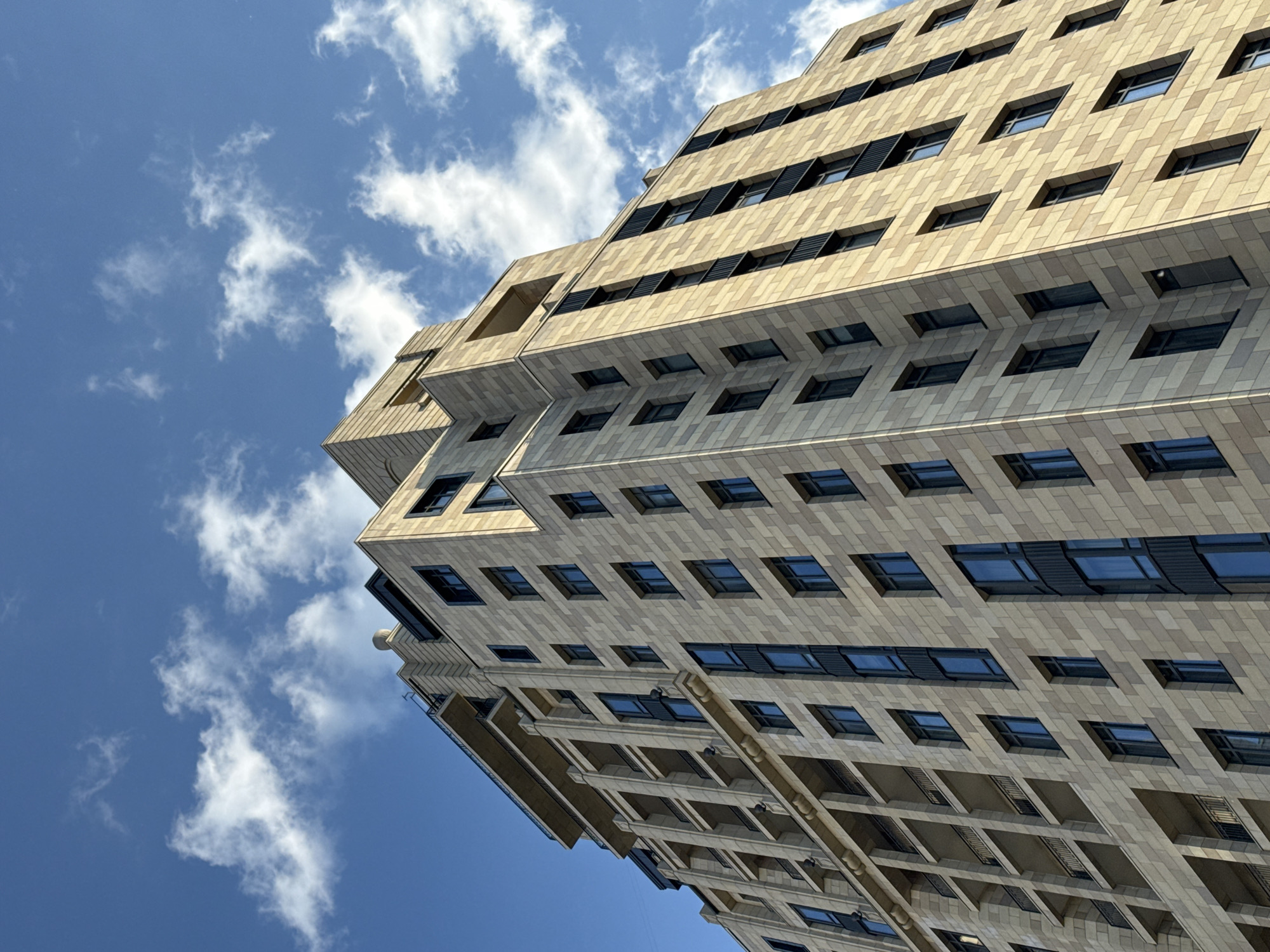 A modern, multi-story building dominates the frame, captured from a low-angle perspective looking upwards towards the sky. The building's exterior is clad in light-colored, possibly beige or sandstone, rectangular tiles, arranged in a repeating pattern. Its design features a contemporary, geometric aesthetic with stepped or tiered sections creating a dynamic silhouette.
Numerous rectangular windows, framed in dark material, are visible across the façade. Some windows appear to have dark internal blinds or external louvers. The building exhibits a mix of deeply recessed windows and more surface-aligned ones, adding to its textured appearance. Balconies or other protruding/recessed architectural elements cast subtle shadows, hinting at the strong sunlight.
The backdrop is a vibrant blue sky dotted with scattered, fluffy white clouds, indicating fair weather. The bright illumination suggests it is daytime, likely mid-day, given the clear visibility and distinct shadows. No people or specific activities are depicted, focusing entirely on the architectural structure and the sky. No text is visible anywhere in the image. The scene presents a clean, modern urban landscape with a strong emphasis on architectural lines and the expansive sky.
