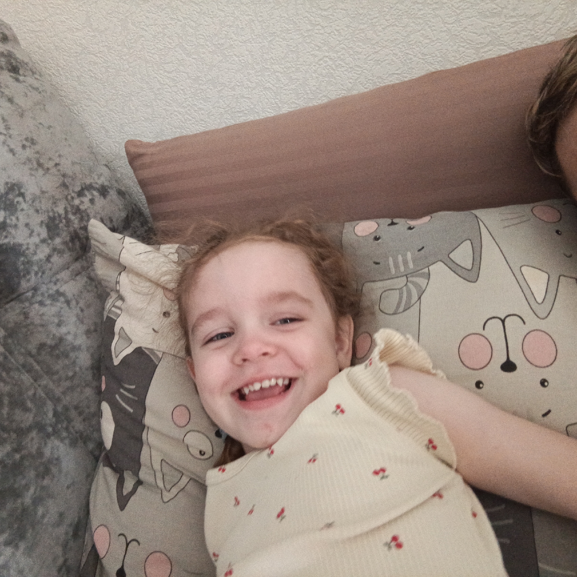 A young child with light reddish-brown hair is lying on a pillow with cat prints. The child is smiling and wearing a light-colored, possibly ribbed, shirt with small red cherry patterns. The pillow is on a couch with a gray velvet-like texture. There is another pillow above it that is brown. The location is Марковский, Russia.