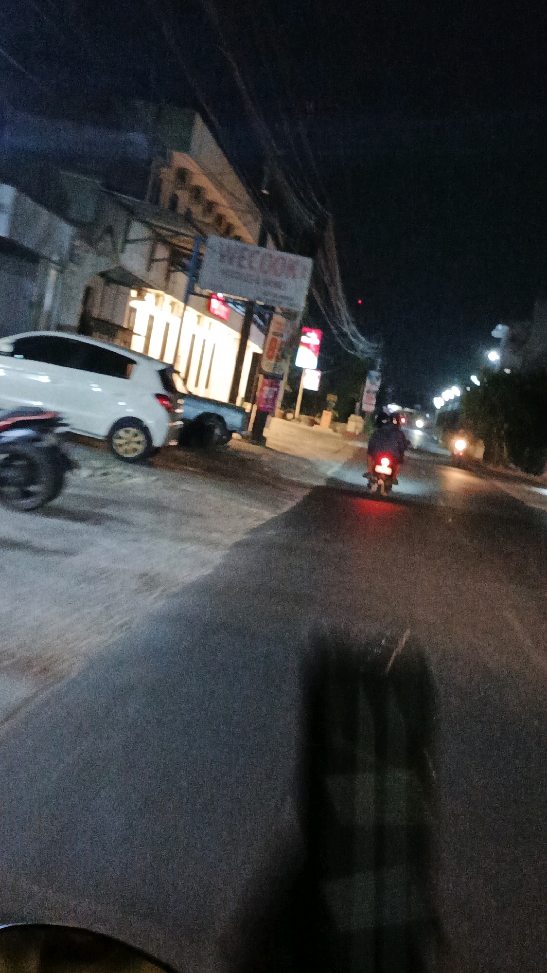 A blurry, nighttime photograph depicts a street scene in Blulukan, Indonesia. A white hatchback is parked on the left side of the asphalt road, partially obscuring a motorcycle's front wheel. Further down the road, a person on a motorcycle with illuminated red taillights is moving away from the camera. On the right side of the road, a building with lit windows is visible, and a sign with the text "WE BOOK" is mounted above it. Power lines crisscross the dark sky. The overall image quality is low, with significant noise and motion blur, suggesting it was taken with a handheld device in low-light conditions.