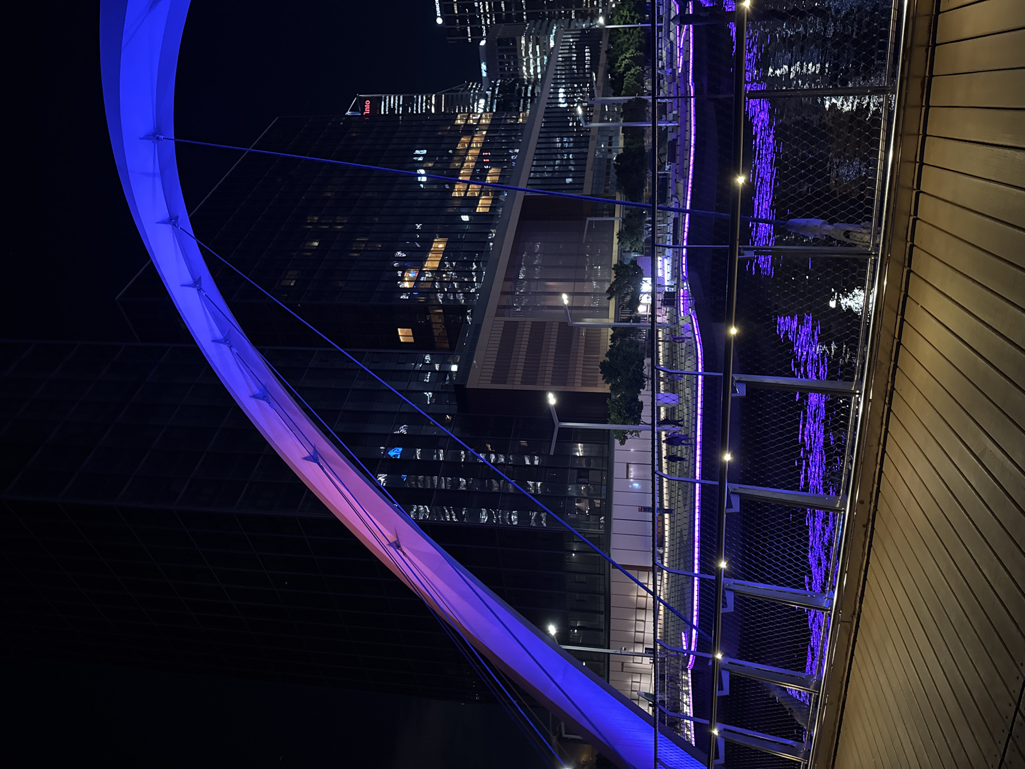 The image displays a striking night scene of a modern city, most likely Perth, Australia, centered around an illuminated pedestrian bridge. The prominent feature on the left is a contemporary bridge with a graceful, curved arch structure that emits a vibrant blue and purple glow. Slender cables extend from the arch, anchoring the bridge deck.
To the right, a wooden-paneled walkway or wall is visible in the foreground. Adjacent to it is a metal railing, interspersed with small, warm-toned lights, overlooking a body of water. The dark water beautifully reflects the purple light from the bridge and surrounding city, creating shimmering patterns.
In the background, a collection of tall, modern city buildings forms the skyline. Numerous windows in these skyscrapers are brightly lit, indicating urban activity. On one building, the word "Inio" is clearly visible in white text, with the "STARBUCKS" logo also faintly discernible above it. Scattered trees are visible near the base of some buildings, adding a touch of nature.
The scene is set at night, with the deep darkness contrasted by the profusion of artificial lights. The weather appears clear. No people are visible, allowing the focus to remain on the architectural beauty and the dynamic urban environment. The image evokes a sense of modern serenity and the captivating allure of a city after dark.