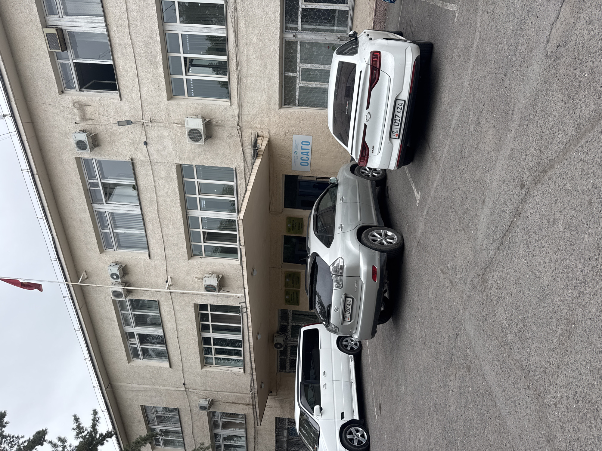 An overcast day captures a frontal exterior view of a modest, light-colored building, likely an office or institutional structure, with a gray asphalt parking lot in the foreground. The building, estimated to be two to three stories high, features numerous windows with white frames, some of which are partially or fully open. Several air conditioning units are visible mounted on the exterior walls. A red flag flies from a flagpole on the roof.
In the parking lot, three vehicles are present. On the left, a white minivan, possibly a Honda Stepwgn, is parked parallel to the building. To its right, a silver Lexus RX SUV is parked, facing away from the camera. To the right of the SUV, a white sedan, identified by its license plate as registered in Kyrgyzstan (KG 017 AZA), is parked, also facing away. A sign above the entrance to the building is visible with Cyrillic text and the abbreviation "ОСАГО" in blue letters. Based on the sign and the visual cues, this location is likely the "OAGAG" office in the city of Kara-Balta, Kyrgyzstan. The environment is clean and orderly, with no visible people in the frame.