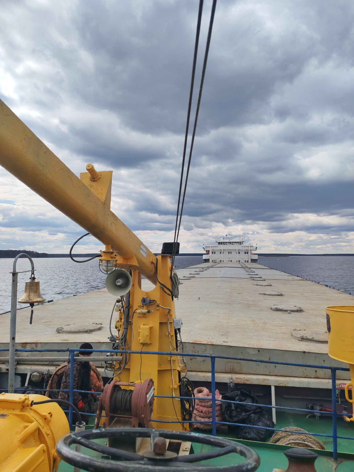 The image presents a working view from the deck of a vessel on a wide body of water, likely a river or large lake in the vicinity of Городище, Russia. The sky is heavily overcast with dark grey clouds, suggesting a cloudy day, but ample light indicates daytime.
In the foreground, a prominent bright yellow crane or boom arm extends from the left, fitted with a megaphone-like speaker, a light, and various electrical connections. A ship's bell hangs from a curved metal post on the far left. At the bottom center, a dark, weathered steering wheel is visible, while to its right, a red-brown winch with coiled rope suggests maritime operations. Blue railings separate the immediate foreground from the main deck. Behind these railings, partially obscured, a person wearing a dark beanie, a camouflage-patterned jacket, and orange gloves is seen, appearing to be working or observing. Stored below the deck level are pinkish coiled ropes and black garbage bags.
The main deck, which is wide and flat with several circular hatches, stretches towards the distant horizon. In the background, a large white multi-deck structure, resembling the bridge and superstructure of another vessel or a prominent section of a large barge, is clearly visible. The water extends to a distant landmass or treeline on both sides, reinforcing the inland waterway setting.
A white label with the text "P9350320" is visible on the yellow crane arm. A red sign, partially seen near the right edge on a yellow structure, appears to read "ПП". The overall scene captures a functional maritime environment, likely involving cargo transport or similar operations, under a characteristically cloudy sky.