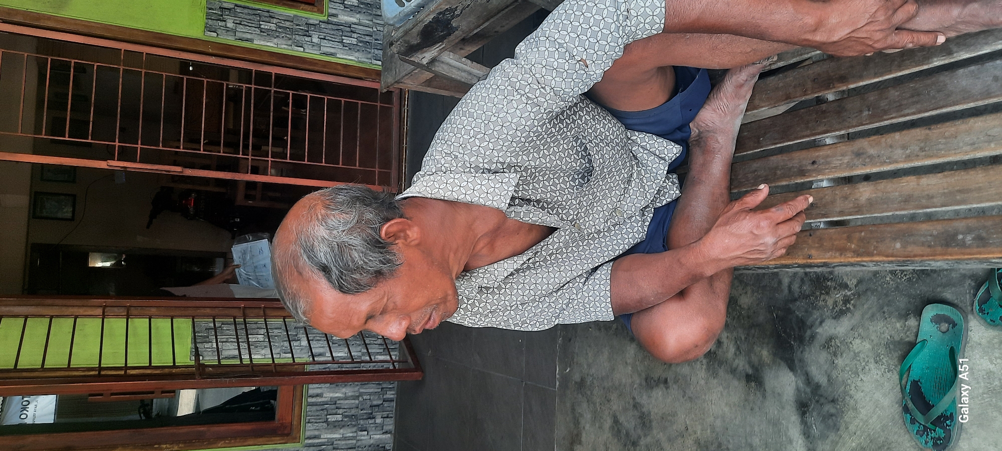 An elderly Indonesian man with thinning gray hair and weathered skin sits cross-legged on a wooden bench in front of a building. He is wearing a short-sleeved collared shirt with a patterned design and dark blue shorts. His feet are bare. A pair of worn green flip-flops are visible on the gray concrete ground to his left. The background shows a doorway with a metal grate, revealing the interior of a room where a motorcycle and some papers are visible. The building appears to be located in a residential or commercial area of Selopuro, Indonesia, with a patterned tile wall and a wooden door frame visible to the left of the man.