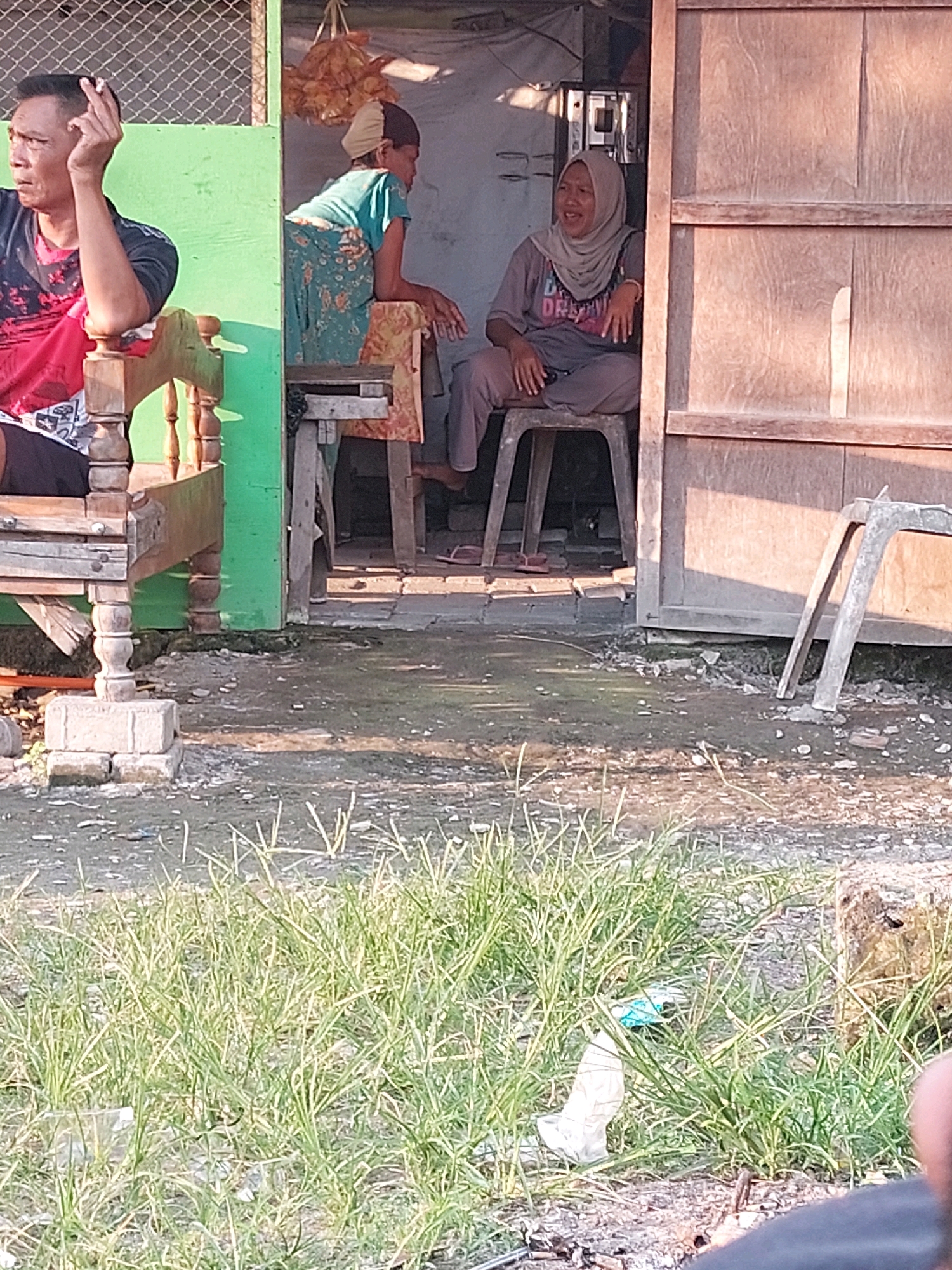 In Canga'an, Indonesia, three individuals are observed in an outdoor setting, likely a roadside stall or a small dwelling. On the left, a man with short black hair sits on a wooden chair, wearing a red and black t-shirt and dark shorts, with a cigarette in his right hand. To his right, inside an open structure, a woman wearing a blue t-shirt and patterned skirt is leaning on a wooden surface. Further to the right, a woman wearing a light grey hijab and a grey t-shirt with the word "DREAM" printed on it, and grey trousers, sits on a plastic chair, her mouth open as if speaking or laughing. The environment consists of a green wall, wooden structures, and a dirt ground covered with debris and some sparse grass in the foreground. Two empty plastic chairs are propped against a wooden wall on the right side of the image.