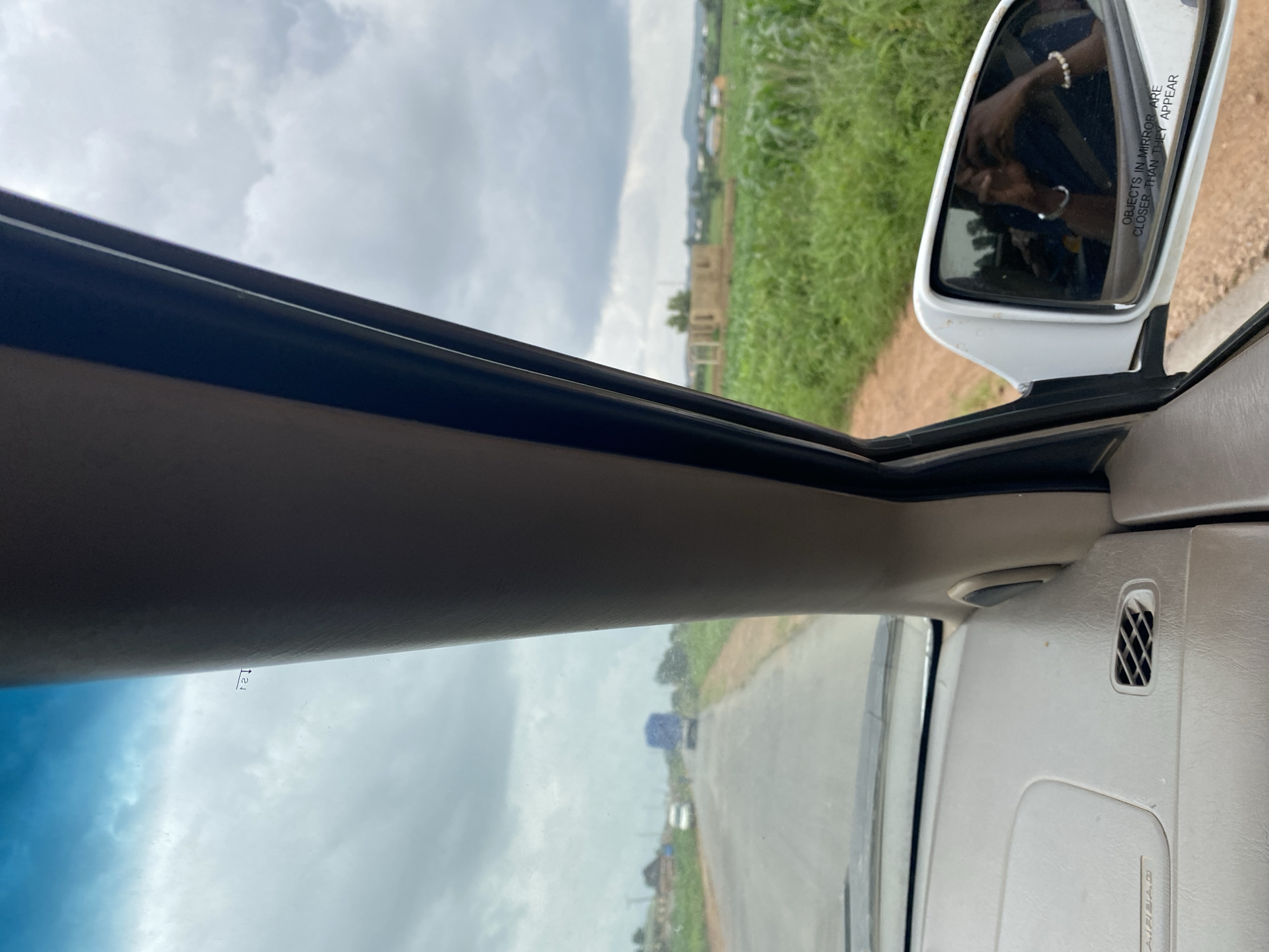The image depicts a view from inside a vehicle traveling on a road in Kuru, Nigeria. The perspective is from the passenger side, looking forward and to the right. On the right side of the frame, a white side mirror is visible, reflecting the interior of the car and a person taking the photograph, presumably wearing jewelry on their wrists. The side mirror has the text "OBJECTS IN MIRROR ARE CLOSER THAN THEY APPEAR."
The interior of the car is partially visible, showing a beige dashboard with an air vent. The windshield displays a cloudy sky. Outside the vehicle, a paved road stretches ahead, with a white van and a blue-covered truck in the distance. To the right of the road, there is a field of green crops, likely maize, and in the background, a partially constructed building and scattered houses can be seen. The overall environment appears rural, under an overcast sky.