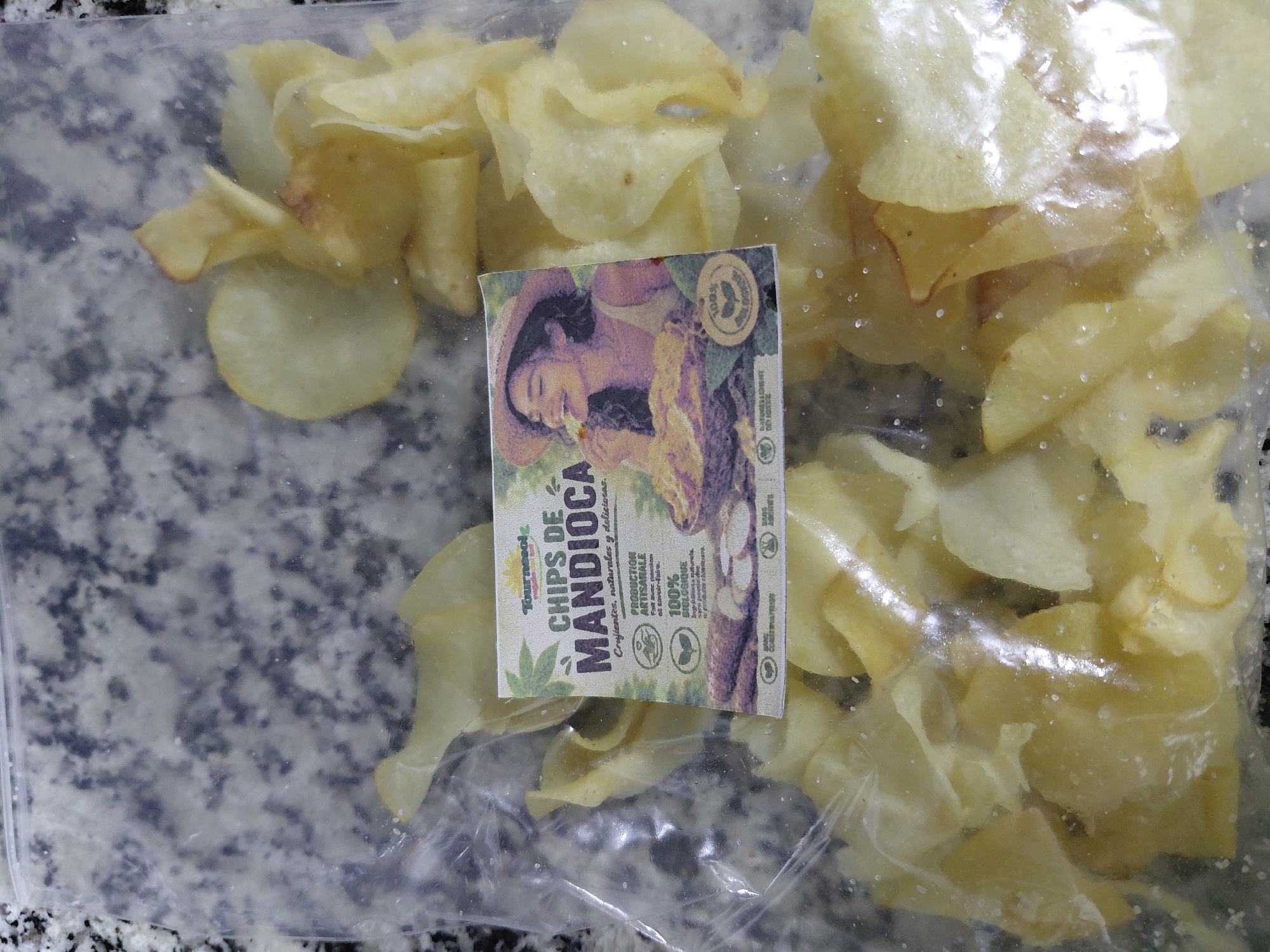 A clear plastic bag filled with cassava chips is displayed on a speckled gray surface, possibly a countertop. A colorful label is affixed to the bag, featuring an illustration of a woman in a straw hat eating a chip, with text in Portuguese indicating "CHIPS DE MANDIOCA" (Cassava Chips), along with claims of being "Crocantes, naturais y deliciosos" (Crunchy, natural, and delicious). The label also highlights "PRODUÇÃO ARTISANAL" (Artisanal Production) and "100% BIOLOGIQUE" (100% Organic). The background of the image is out of focus, showing a mottled gray texture consistent with a granite or marble surface. No specific location in Encarnación, Paraguay, is identifiable from the visual information provided in the image.