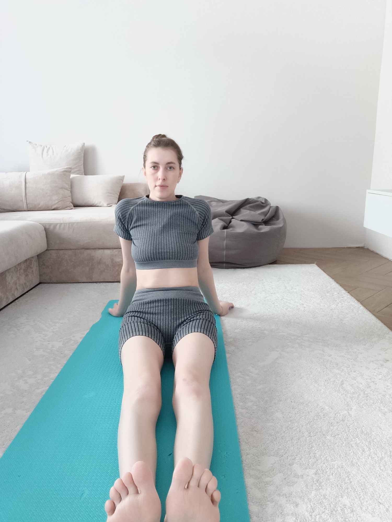 A woman sits on a blue yoga mat in a room with a white wall, beige sofa, and gray beanbag chair. She is wearing a gray striped crop top and shorts, and her legs are extended straight in front of her. Her bare feet are visible in the foreground. The room appears to be a living room or home workout space. No information related to the city of Chaikovsky, Russia, is discernible from the image.