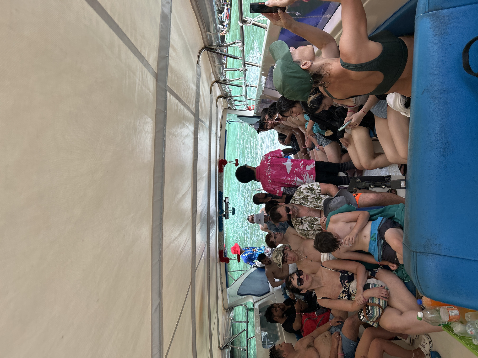 The image shows a crowded boat with many people on it, traveling through the turquoise waters of Ao Nang, Thailand. The boat has a white canopy providing shade, and blue seats are visible. Passengers are dressed casually in swimwear and beach attire. Some people are looking at their phones, while others are talking or observing the surroundings. The water appears calm and clear, with other boats visible in the background, suggesting a popular tourist area. The overall atmosphere seems relaxed, typical of a day trip on the water in a tropical destination.