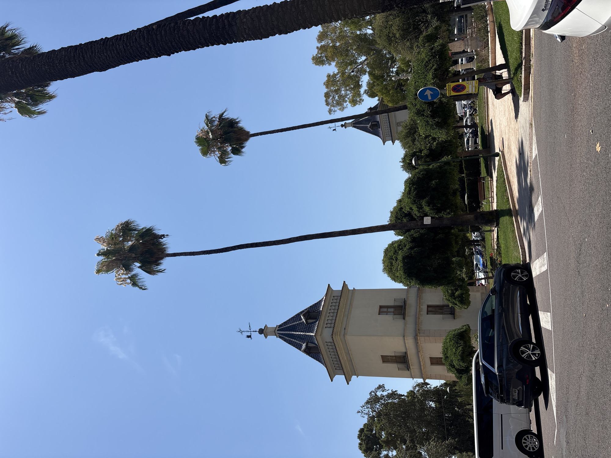 The image depicts a bright, sunny day in Valencia, Spain, with a clear blue sky. The scene is an outdoor urban environment, likely a street or park area, characterized by lush green trees and palm trees. A prominent architectural feature is a light-colored building with a distinctive dark tiled, pointed roof, possibly a church tower or a historical building.
In the foreground, a dark-colored car is parked on the right side of the asphalt road, partially obscuring a white van. The road has white markings. To the left of the road is a paved walkway bordered by a grassy area. A person, seen from behind, is crouched down near the edge of the walkway, wearing a checkered shirt and dark shorts. They appear to be interacting with something on the ground or looking at something on the walkway. Several traffic signs are visible on posts along the walkway, including a blue circular sign with a white arrow pointing up and a yellow rectangular sign with red markings, suggesting parking regulations.
Several tall palm trees are a significant element of the scene, with their fronds blowing gently, indicating a light breeze. One large palm tree trunk dominates the upper left portion of the frame, stretching diagonally across the sky. Other palm trees are seen further down, framing the building and the sky. The time of day appears to be midday or early afternoon, given the strong sunlight and the shadows cast. The overall mood is serene and typical of a warm, pleasant day in a Mediterranean city.