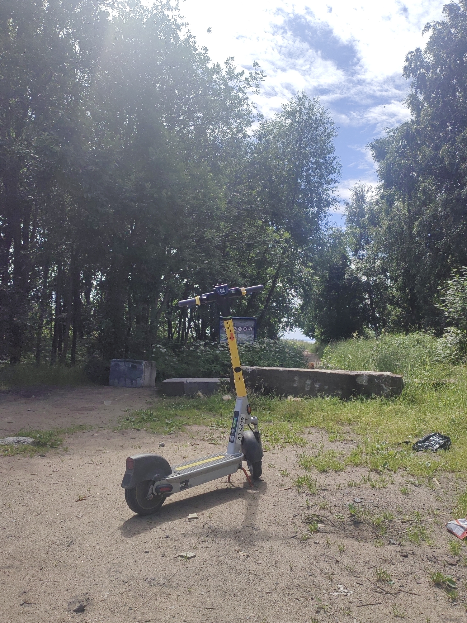 A yellow and grey electric scooter is parked on a sandy path in an outdoor, wooded area. The scooter is facing left and is positioned in the foreground. Behind the scooter, a line of trees with dense green foliage stretches across the background. The sky above is blue with scattered white clouds. In the mid-ground, there are some low concrete structures and overgrown grass. The lighting suggests it is a sunny day with some glare from the sun. The environment appears to be a park or a natural area on the outskirts of a city, possibly in Saint Petersburg, Russia, given the context of similar images.