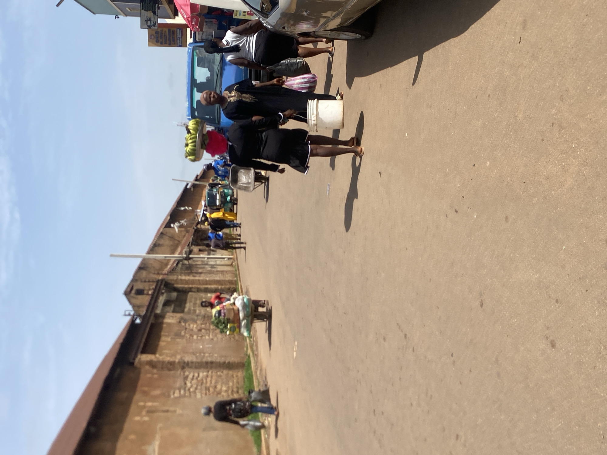 In Bukuru, Nigeria, a dusty street is lined with brick buildings on the left and businesses on the right. Several people are present: a woman in a black dress carries a bucket and bags, and another woman in a black outfit walks ahead, wearing a red headscarf and a tray of green fruit on her head. A man in a red shirt pushes a wheelbarrow laden with produce, while other individuals walk in the background, some carrying bags. A blue truck is parked on the street, and a silver car is visible on the right. The scene captures a bustling market atmosphere with everyday activities.