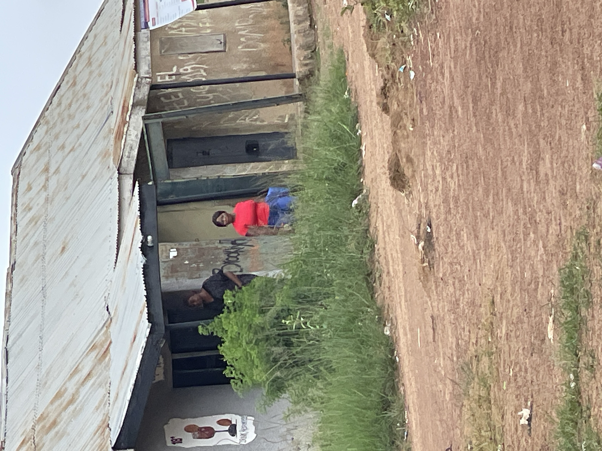 The image depicts an outdoor scene in Durumi, Nigeria. The primary subjects are two individuals. One person, wearing a red shirt and blue bottoms, stands in front of a building. Another person, dressed in a dark shirt, is visible in the background, partially obscured by greenery. The building appears to be a simple structure with a corrugated metal roof and possibly concrete walls. There is graffiti on the building's exterior.
The scene is situated alongside a dirt path or road, with an area of dense green vegetation growing between the path and the building. The ground is dry and appears to be earth or sand, with scattered debris, suggesting a somewhat unkempt environment. The lighting suggests it is daytime, though the weather is not clearly discernible. The overall impression is of a casually observed moment in a local, possibly semi-rural or developing urban, setting.
The image is oriented vertically, meaning the content is displayed sideways. Some visible text, likely graffiti, is present on the building, but it is illegible due to image quality and resolution. There are no obvious indications of a specific event taking place; the individuals appear to be casually present in the scene.