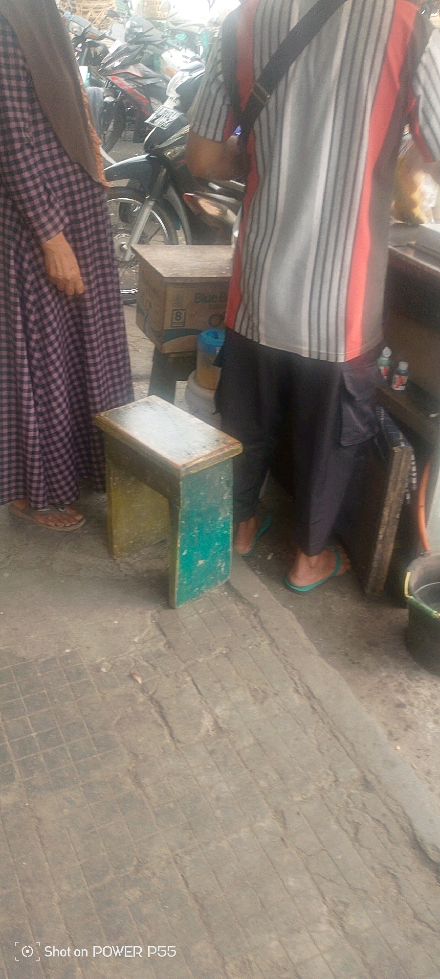 A person wearing a purple and black checkered dress and a brown hijab stands to the left of the frame. To their right, another person wearing a striped shirt and black pants stands facing away from the camera. In the foreground, a small, weathered stool with a green and yellow painted finish sits on a tiled surface. Behind the stool, a cardboard box with the "Blue D" logo and the number "8" is visible, along with a blue-capped container. In the background, several motorcycles are parked. The scene appears to be outdoors in an urban environment.