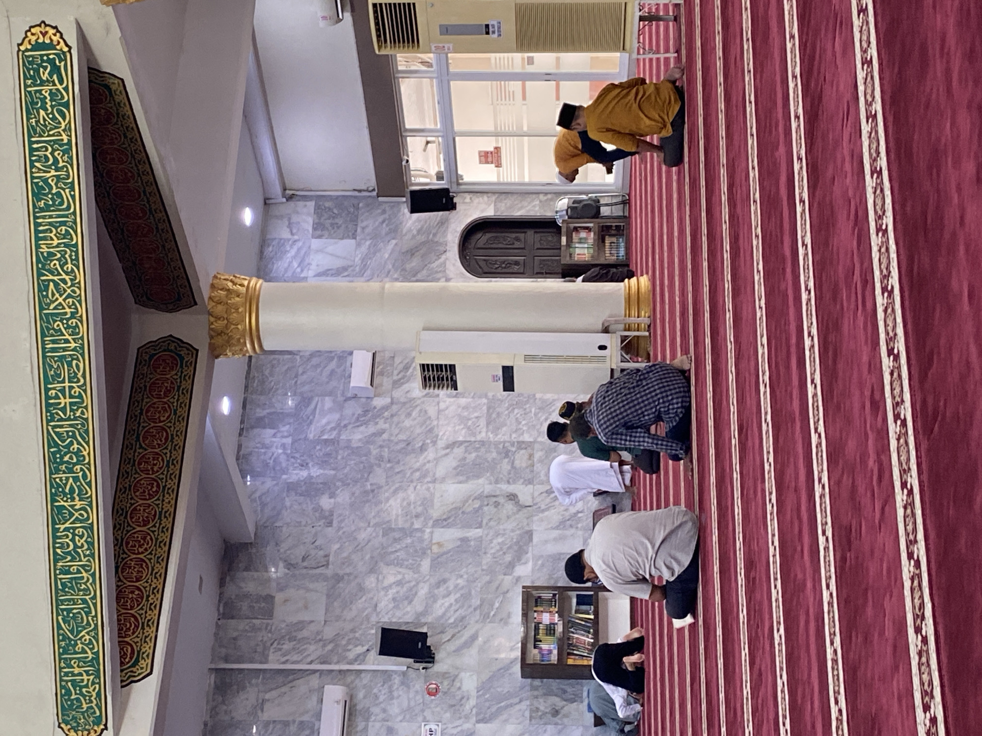In the city of Lhokseumawe, Indonesia, inside a mosque, several men are engaged in prayer. The mosque features a red carpet with white striped borders, a marble-tiled wall, and a large white column adorned with gold accents. Decorative Arabic calligraphy is visible along the ceiling. A bookshelf filled with religious texts is situated against the wall. Several individuals are in various prayer postures, including bowing and sitting. One man in a white robe is standing, and another in a yellow shirt is in a kneeling position. Air conditioning units are mounted on the walls. The lighting is subdued, creating a tranquil atmosphere.