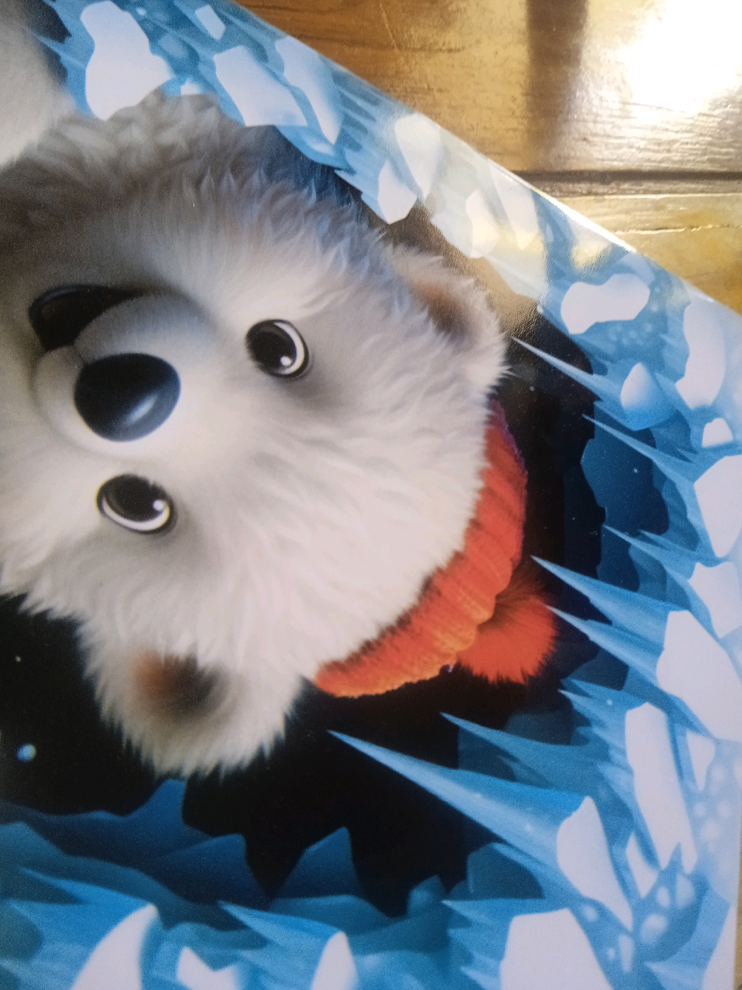 The image shows a close-up of a fluffy white polar bear cub wearing an orange scarf, peeking out from an opening surrounded by blue ice formations. The cub's face is the primary focus, with large, dark eyes and a slightly tilted head. The background is dark, with the blue ice appearing sharp and angular. There is a wooden surface visible in the top right corner of the image, which might be a table or a floor. The image does not contain any information about the city of Probolinggo, Indonesia.