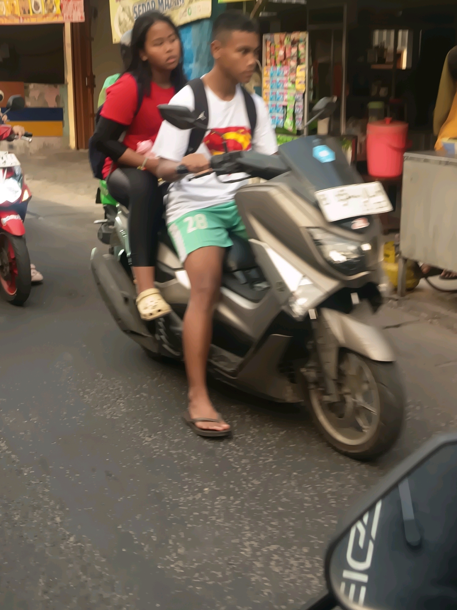 Two individuals, a male and a female, are riding on a silver scooter in the city of Tangerang, Indonesia. The female, wearing a red t-shirt and black leggings, is seated behind the male, who is wearing a white t-shirt with a graphic and light green shorts. The scooter is in motion on an asphalt road, with other vehicles and buildings visible in the background. The overall scene depicts everyday transportation within an urban setting.