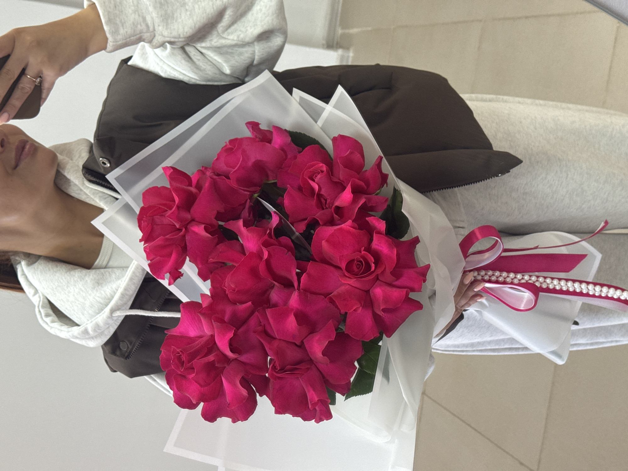 A woman is holding a bouquet of pink roses wrapped in white paper, tied with a pink ribbon and a beaded string. She's wearing a grey hoodie and a brown puffer vest. She's also holding a phone. The location is unknown, and there's no evidence to suggest it's in Chaikovsky, Russia. The environment seems to be indoors.