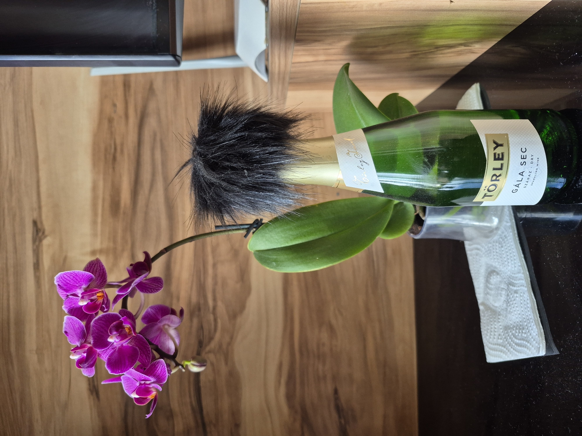 A horizontal, eye-level shot captures a scene in an unspecified location, possibly in Tbilisi, Georgia. The composition is dominated by a wooden surface in the background, with an orchid featuring magenta flowers in the foreground. The orchid is accompanied by a green bottle, possibly a wine bottle, which has a black, furry object attached to its neck. The bottle is surrounded by green leaves. A tissue is also present.