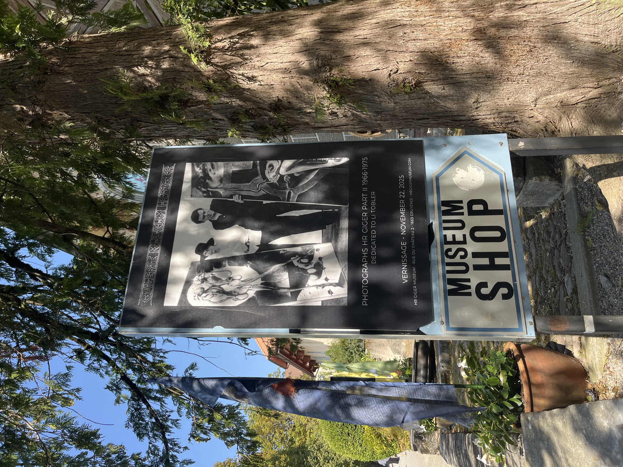 This is a medium, eye-level shot of a large poster and a tree, with a sign for a museum shop in the foreground, on a sunny day. The poster, mounted on a dark backing, features a black and white photograph with the text "GALERIE MUSEUM H.R. GIGER" at the top. Below the photograph, text reads "PHOTOGRAPHS HR GIGER PART II 1966-1975 DEDICATED TO LI TOBLER". Further down, "VERNISSAGE - NOVEMBER 22, 2025" is printed, followed by "HR GIGER MUSEUM RUE DU CHÂTEAU 2 1663 GRUYÈRES HRGIGERMUSEUM.COM".
To the left of the poster, a tall, thin, blue and white sign with an arrow pointing to the right has the word "MUSEUM" in large black letters above "SHOP". The sign is partially obscured by the poster and a tree trunk on the right.
A large, mature tree with rough bark dominates the right side of the frame, casting dappled shadows on the poster and the surrounding area. In the background, a building with a red-tiled roof is visible through the tree branches. To the left of the poster, prayer flags with Tibetan inscriptions are draped, and a potted plant with green leaves sits in a brown terracotta pot in the lower left corner. The ground is paved with cobblestones and gravel. The sky is clear and blue. The location is identified as Epagny, Switzerland, based on the text on the poster indicating "HR GIGER MUSEUM RUE DU CHÂTEAU 2 1663 GRUYÈRES HRGIGERMUSEUM.COM", which is the H.R. Giger Museum in Gruyères, Switzerland. Epagny is a municipality in the canton of Fribourg, near Gruyères.