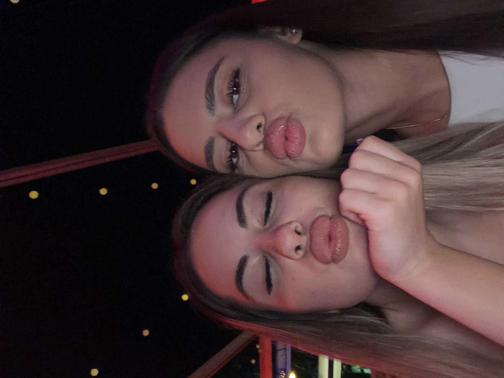 Two young women are posing for a selfie in what appears to be a dimly lit establishment with string lights in the background. The woman on the left has her eyes closed and her lips pursed, with her hand resting on her chin. The woman on the right also has her lips pursed, with her eyes open. Both are looking towards the camera. The image does not provide any specific details that would allow for identification of the location within the city of Haskovo, Bulgaria.