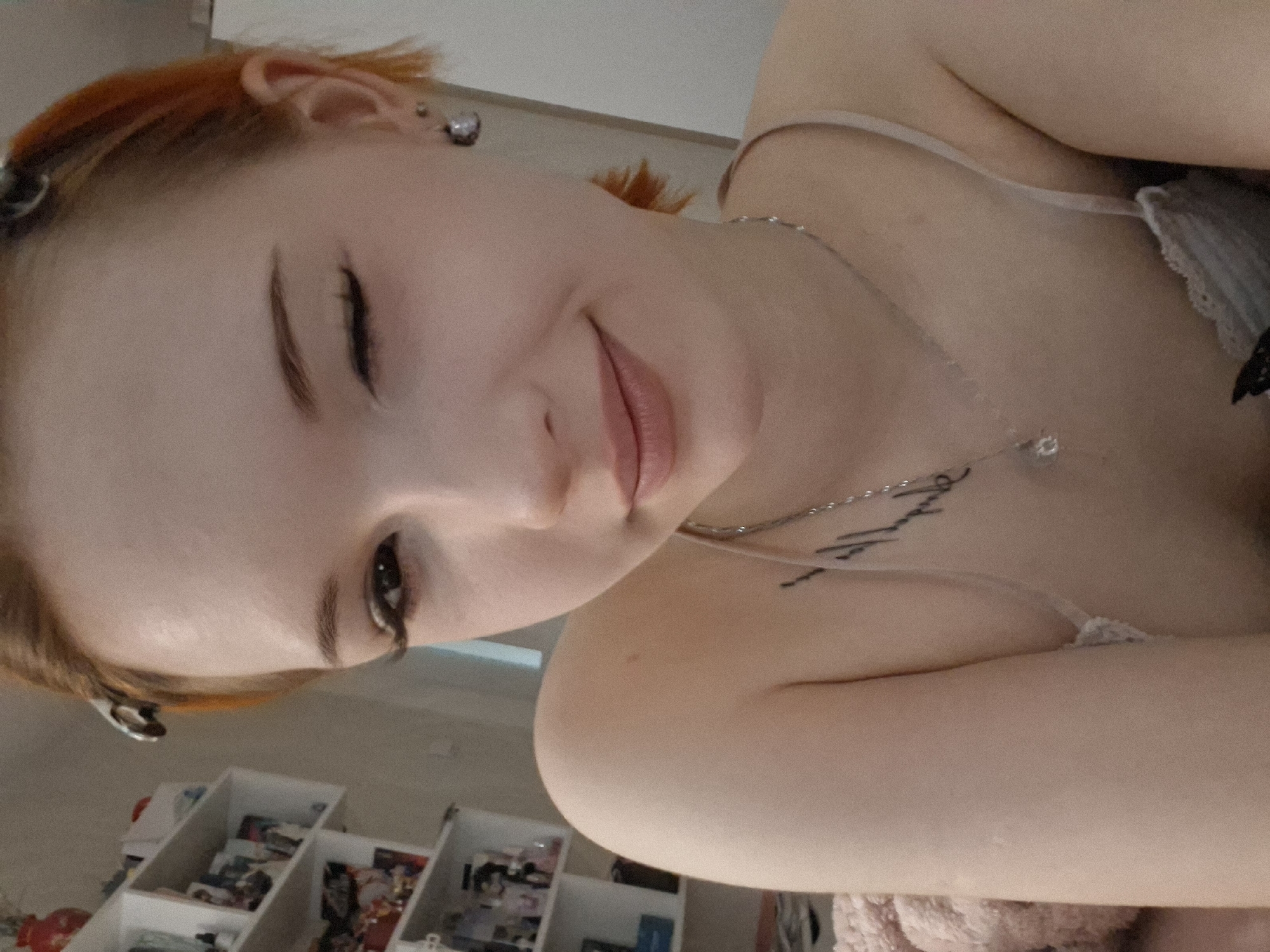 A close-up, indoor shot of a young woman with red hair, pale skin, and a delicate silver necklace lying down. She has a small tattoo on her chest. Her eyes are closed in a smiling expression. A white shelving unit with various items is visible in the background. No evidence suggests the location is Chaikovsky, Russia.